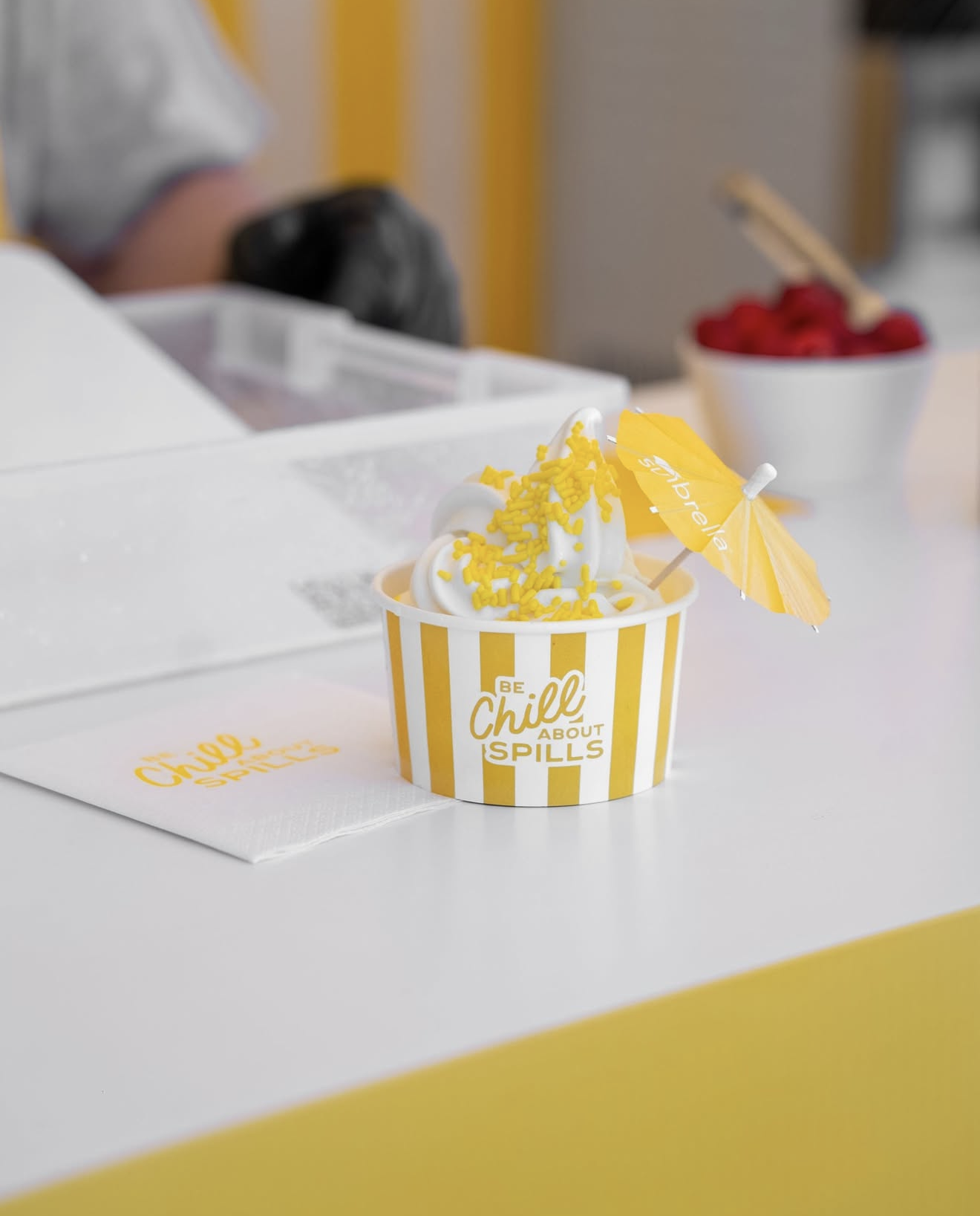 A cup of soft serve ice cream with yellow sprinkles and a yellow paper umbrella, placed on a white napkin on a counter. The cup has yellow and white stripes and the text 'Be Chill About Spills'. In the background, there is a bowl of raspberries and a