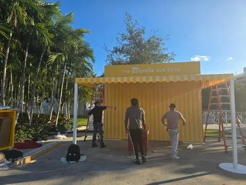 People setting up a yellow sun deck booth with a striped awning, surrounded by palm trees and clear blue sky.