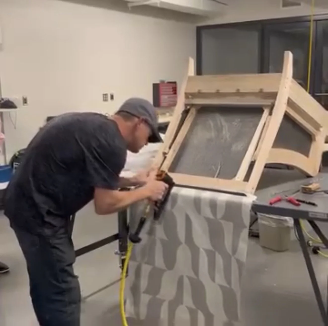 A person working on a piece of furniture with a black and gray patterned cloth draped over it, in a workshop with tools on a table.