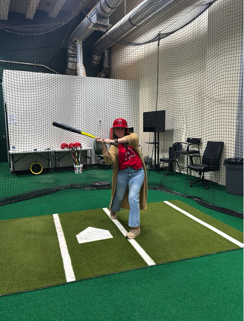 A woman practicing baseball batting in an indoor batting cage, wearing a red helmet and a red Dodgers shirt, holding a bat ready to swing.