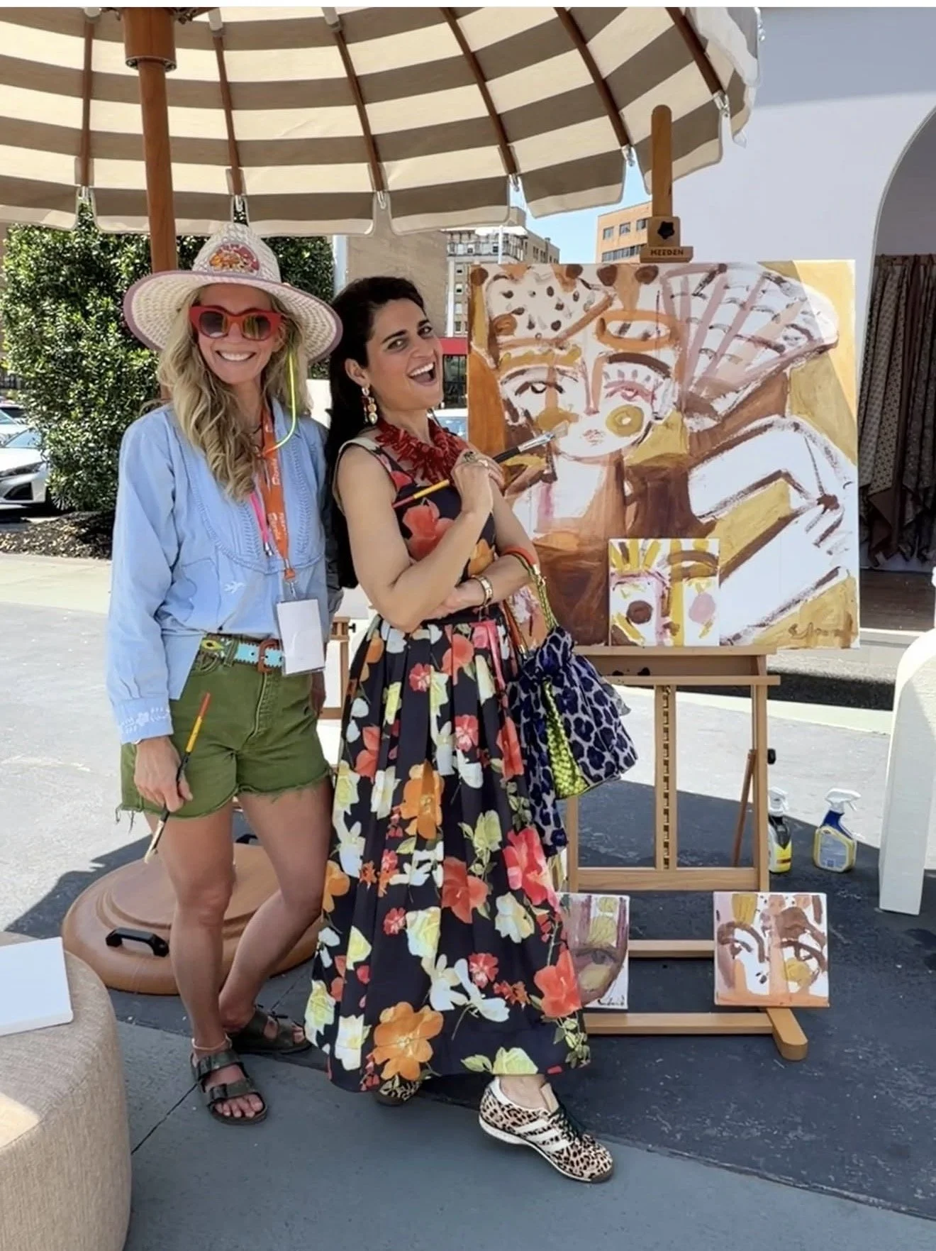 Two women posing next to an abstract painting at an outdoor art event, with one holding a paintbrush, under a striped canopy.
