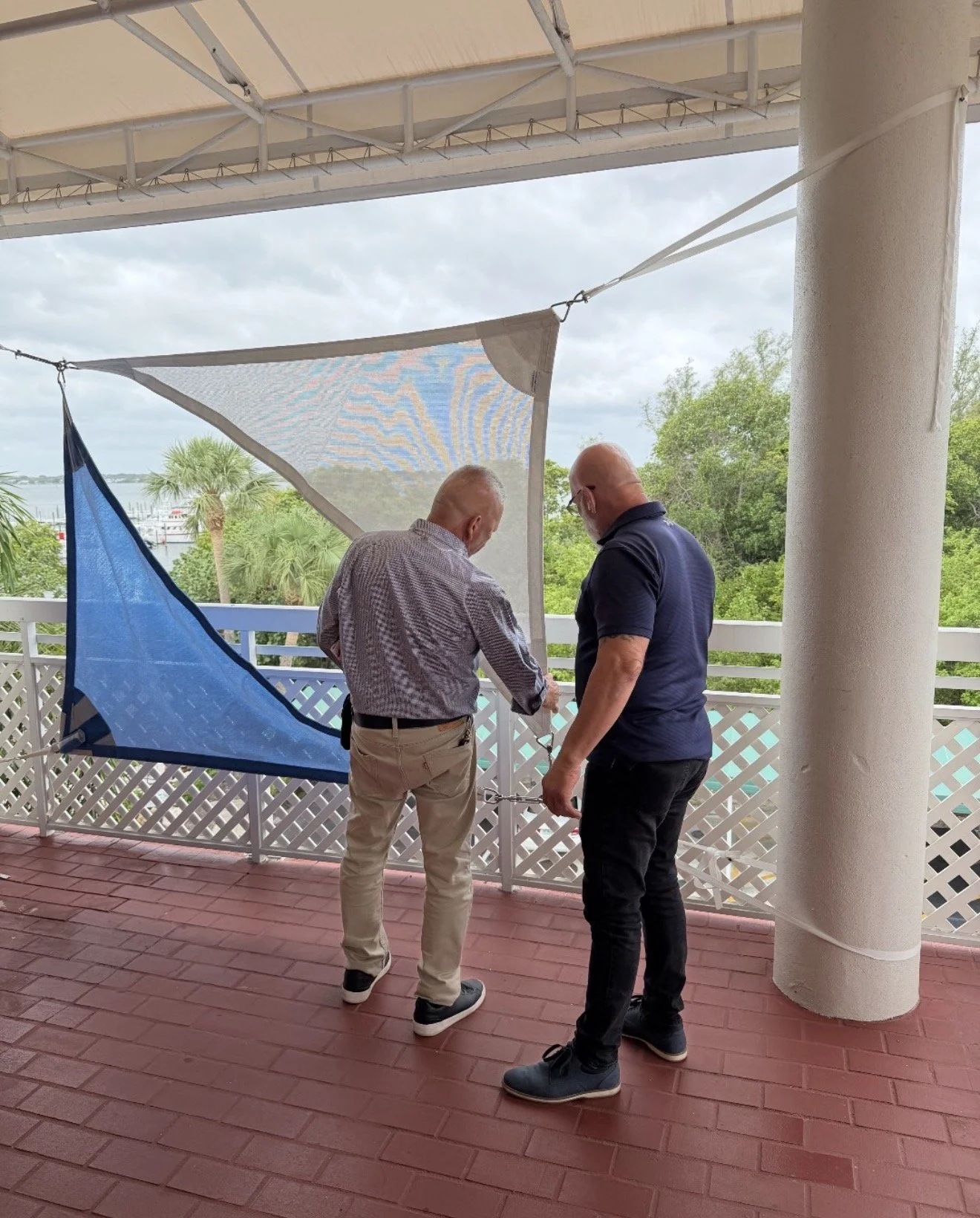 Two men standing on a porch, examining or repairing a safety net or barrier. The porch has a red brick floor and a white railing with lattice design. Outdoors, there are green trees and a body of water in the background, with overcast skies.