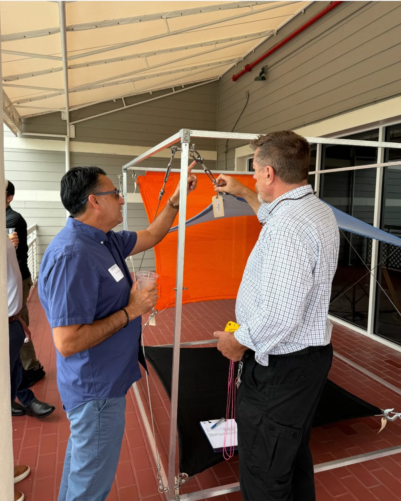 Two men are conducting an experiment with a small weather balloon setup outdoors on a covered patio. One man is in a blue shirt and the other in a checkered shirt. They are attaching equipment under an orange balloon. Several people are visible in the background.