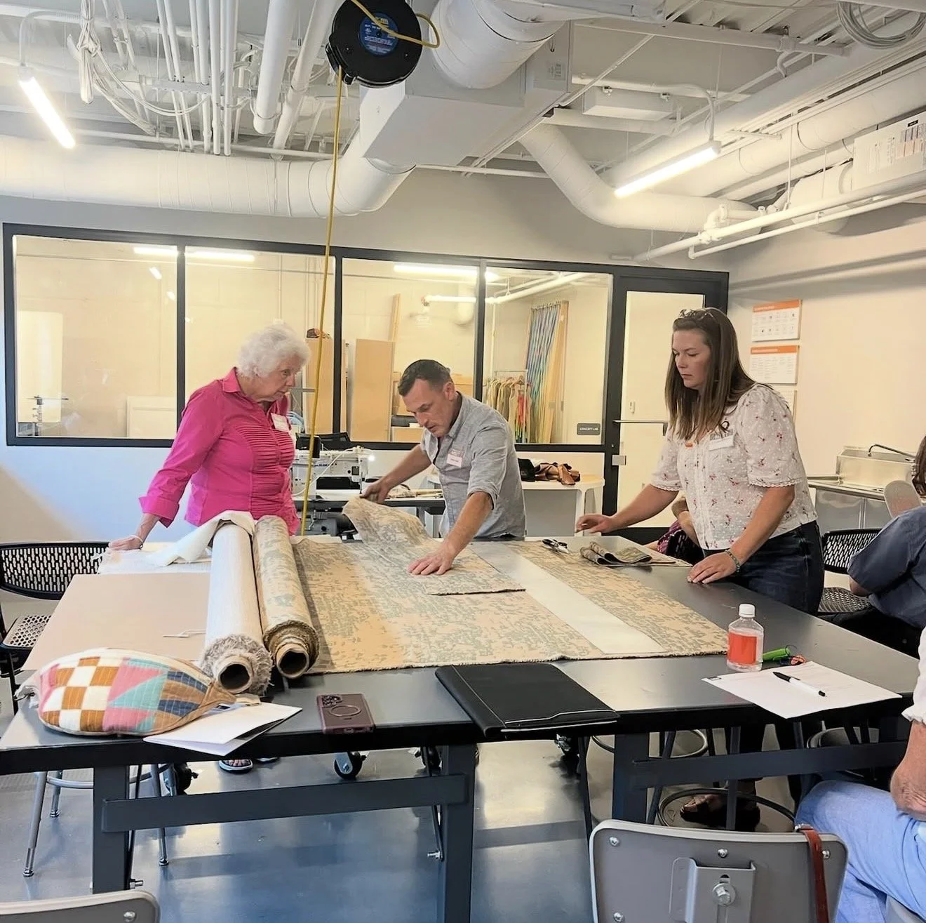 People examining and studying a large rolled-up fabric or rug on a table in a modern industrial-style room.