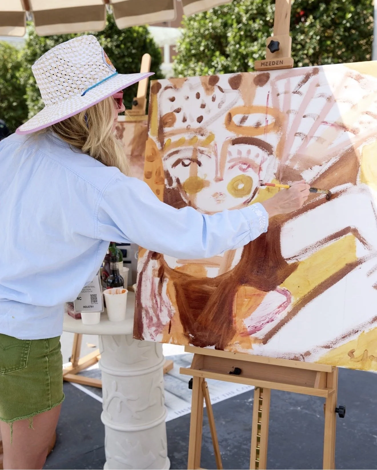 Woman painting an abstract portrait on a large canvas outdoors, wearing a wide-brimmed straw hat and sunglasses, with trees and outdoor furniture in the background.