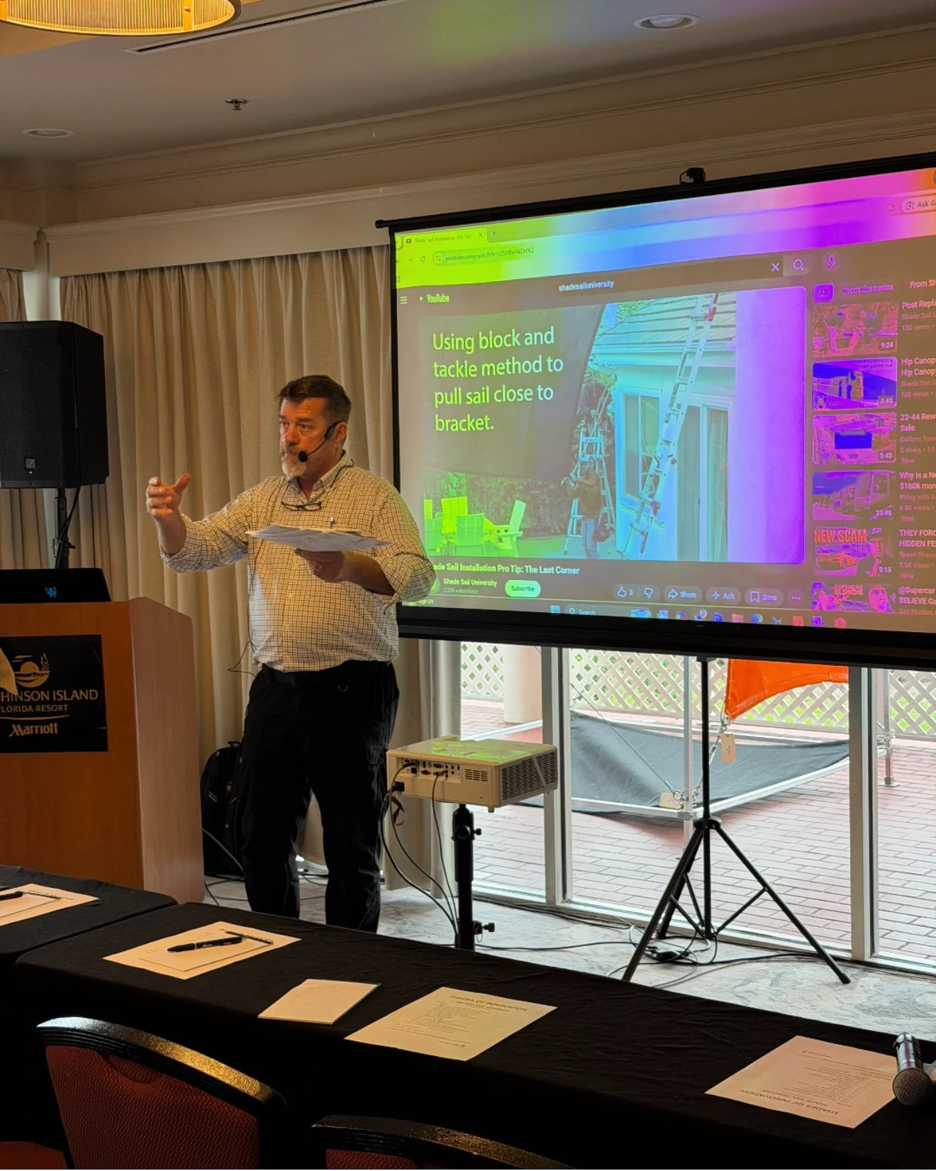 A man giving a presentation in a conference room with a large screen displaying a slide about using block and tackle method to pull sail close to a bracket, with a ladder and some outdoor furniture visible through glass doors in the background.