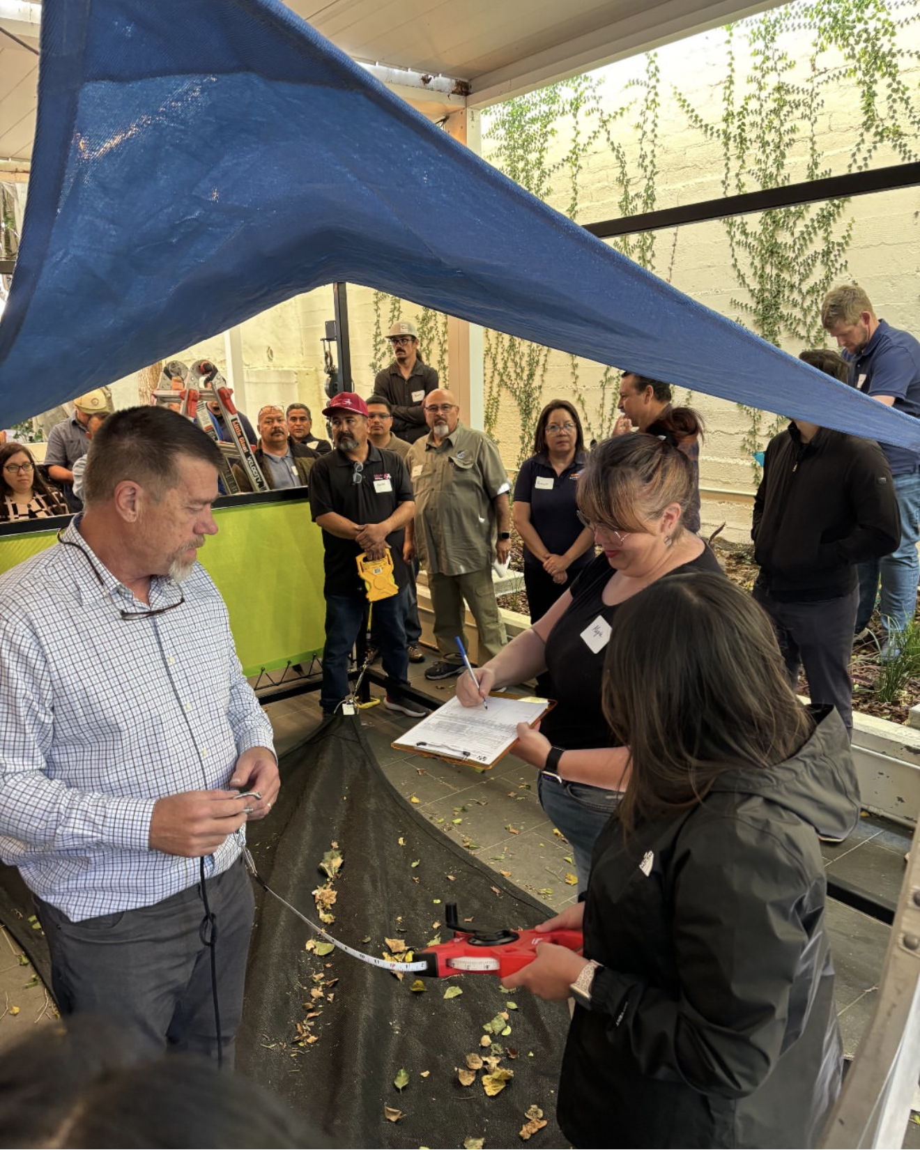 A group of people gathered in what appears to be a workshop or demonstration setting. A man in a checkered shirt holds a measuring tape, and a woman with glasses is writing on a clipboard. Several other people are standing around, some holding tools or equipment. The setting includes a table with leaves on the ground and a large blue canopy overhead, with a yellow-green partition and a wall with creeping vines in the background.