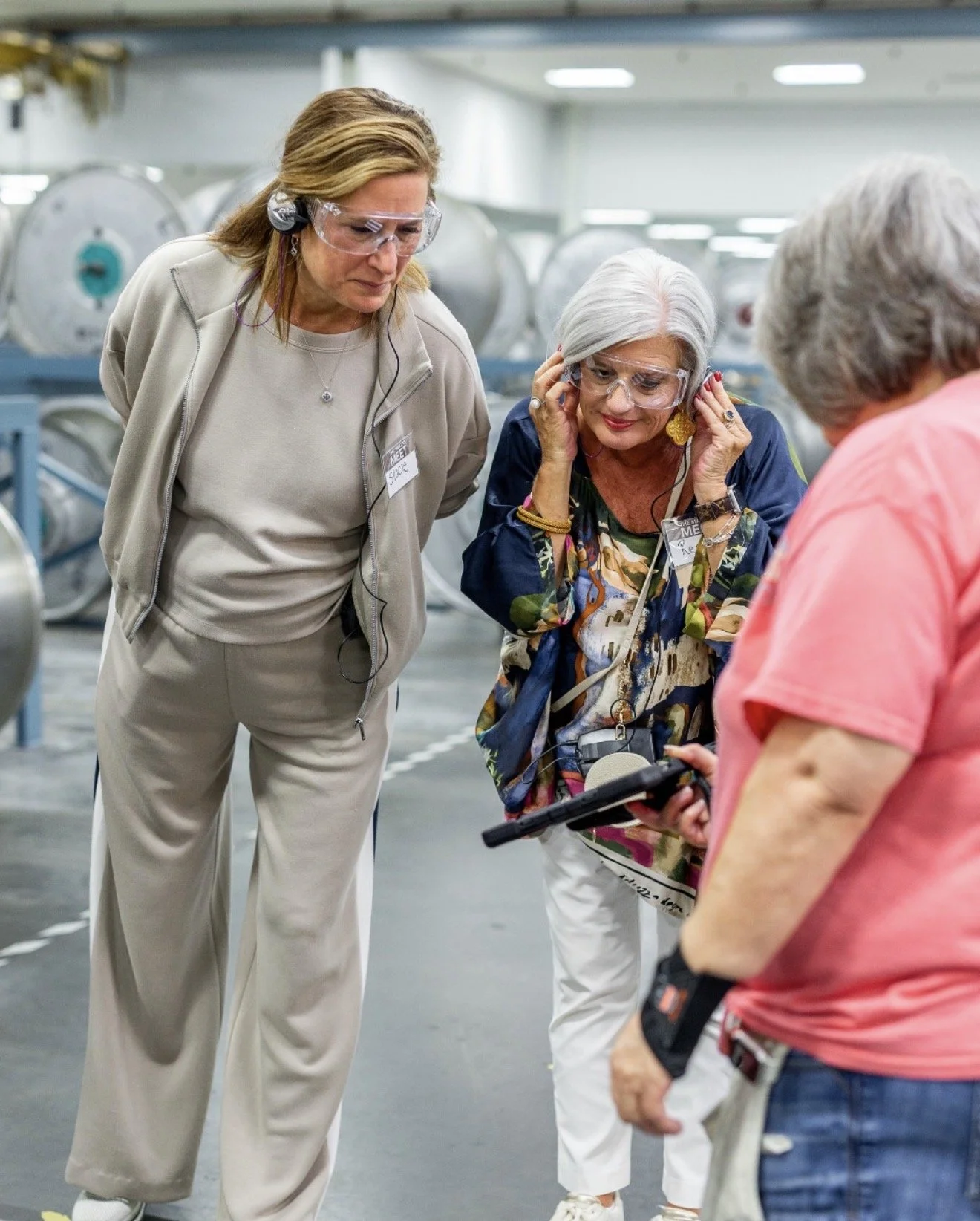 Three women in a factory or warehouse, wearing safety glasses, looking at a device held by one of them, surrounded by large industrial equipment.