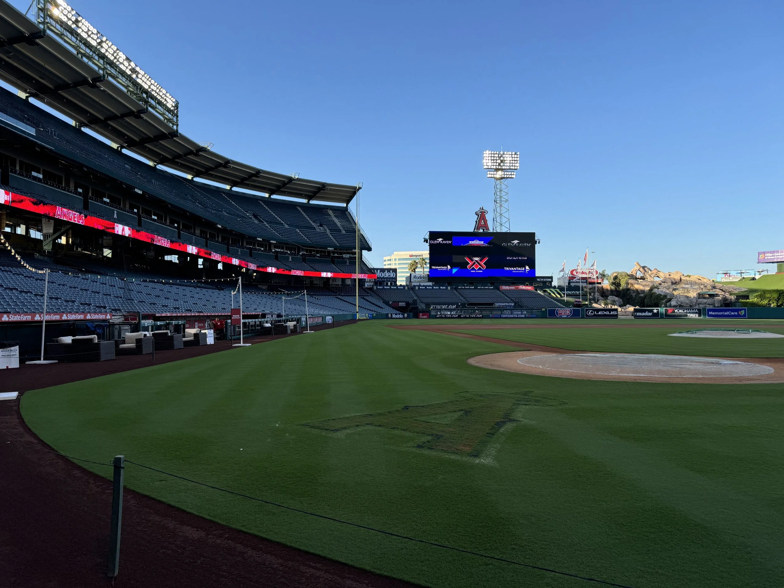 Empty baseball stadium with green field and empty seats, scoreboard, and large lighting towers against a clear blue sky.
