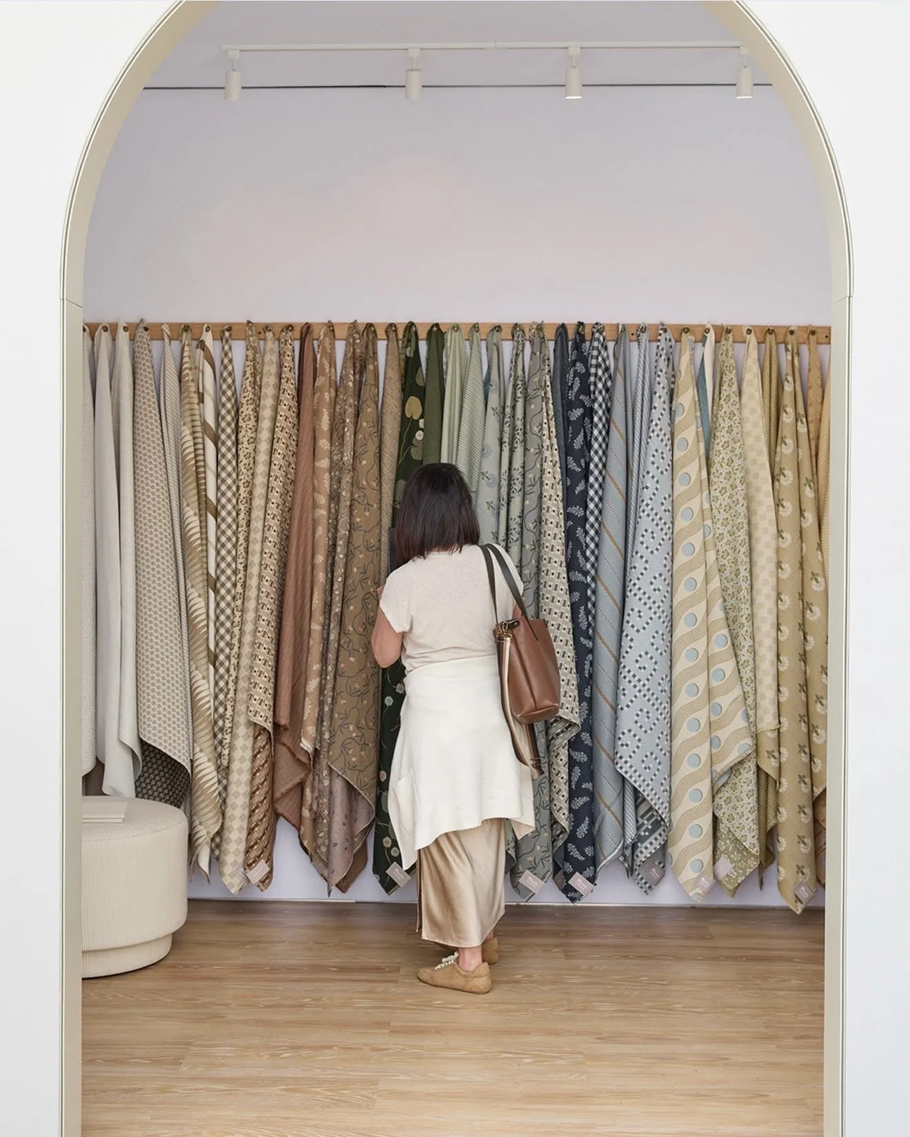 A woman shopping for fabrics in a store, standing in front of a display of patterned fabric rolls on a wall, with a white arched doorway framing the scene.