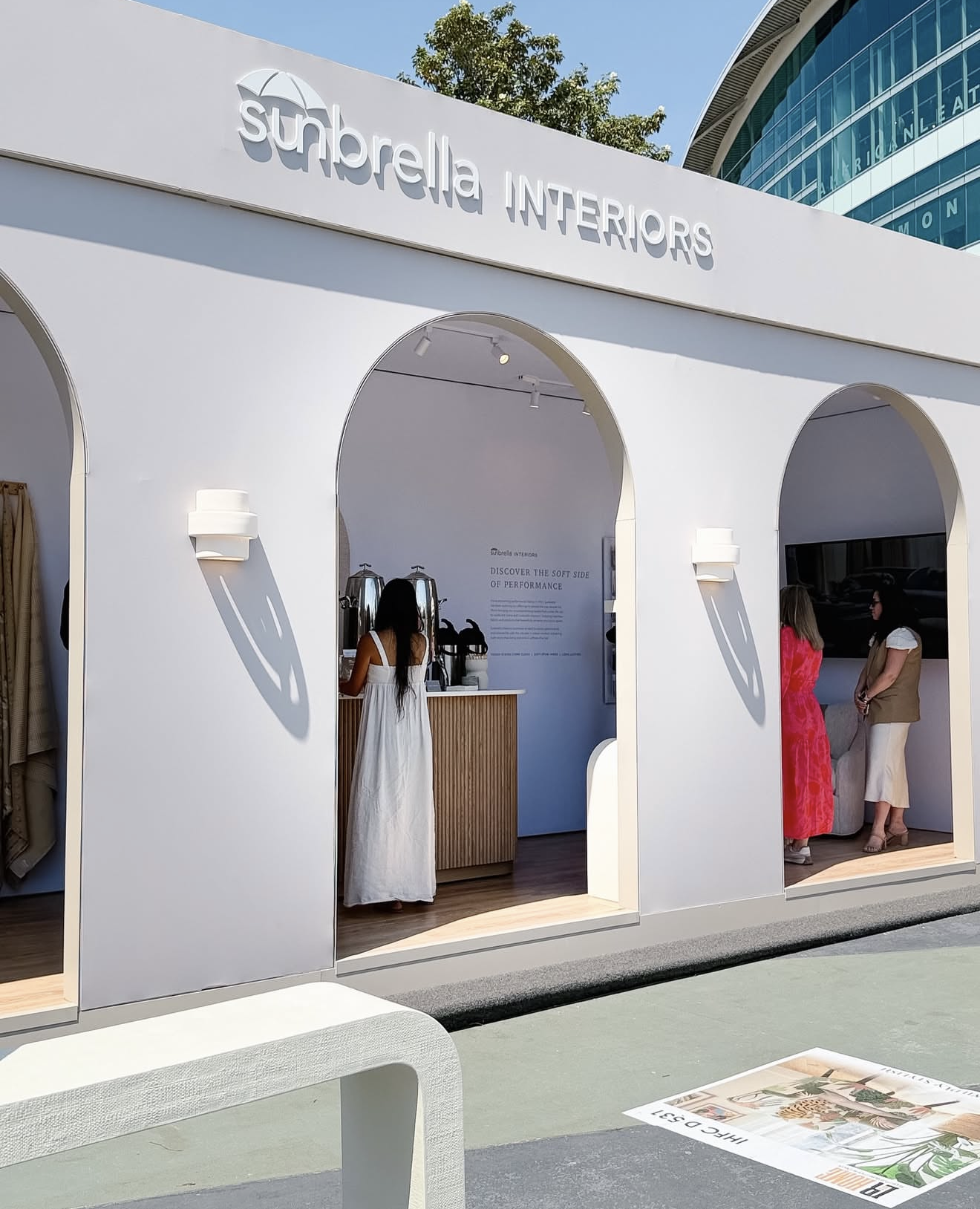 Outdoor booth for Sunbrella Interiors, with three women and a barista, set up under white arches with shadows on the wall, in an urban area with a modern building and trees in the background.