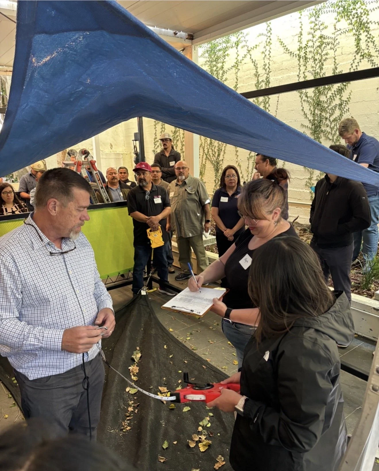 People participating in an indoor workshop or demonstration, with several people observing and one woman writing on a clipboard. There is a blue tarp overhead, and a group of observers, some holding measurement tools, are gathered around a planted ar