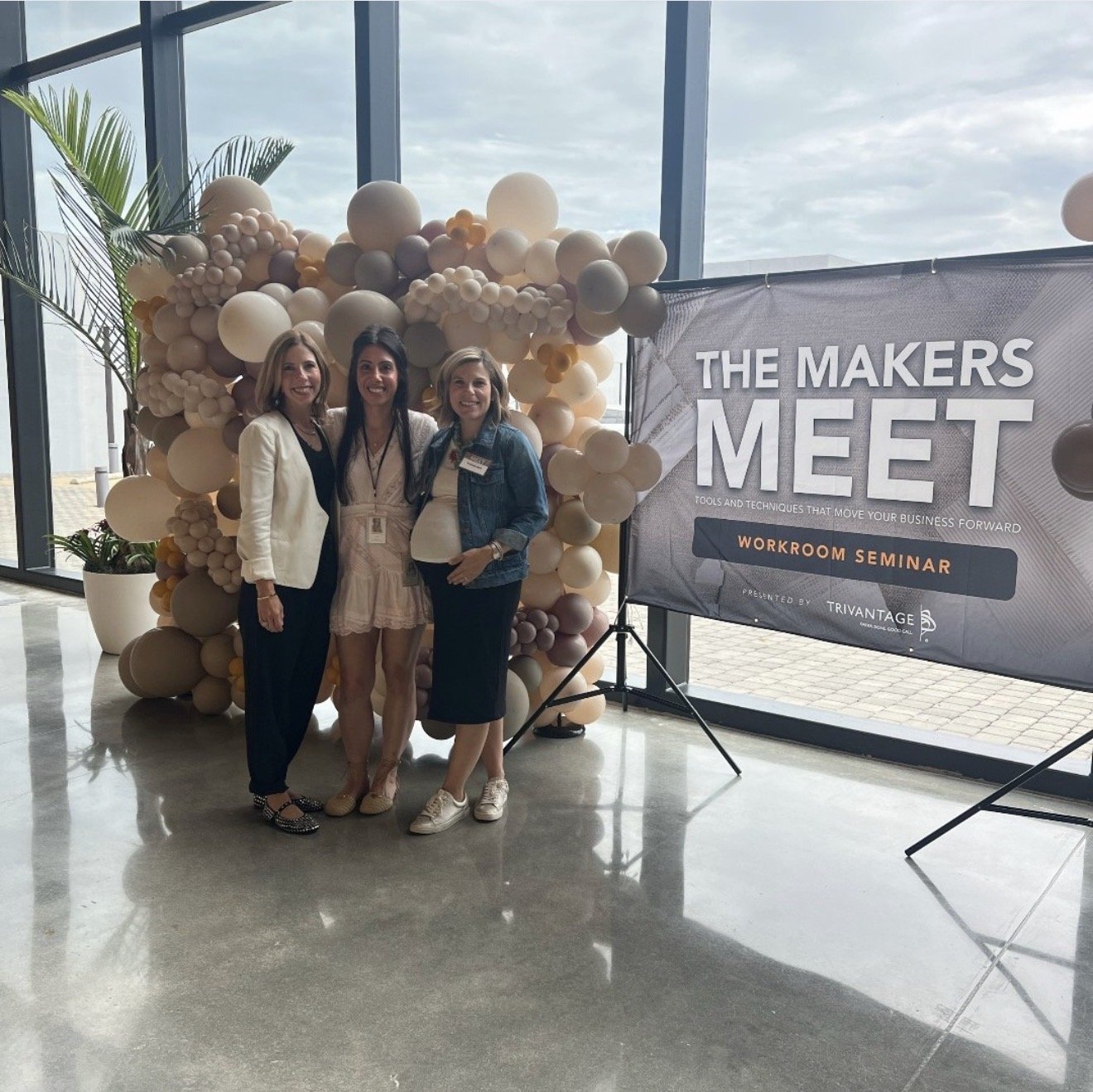 Three women standing together inside a modern building, smiling for a photo. They are near a decorative balloon arch and a large sign that reads 'The Makers Meet', promoting tools and techniques for business success and a workroom seminar, presented 
