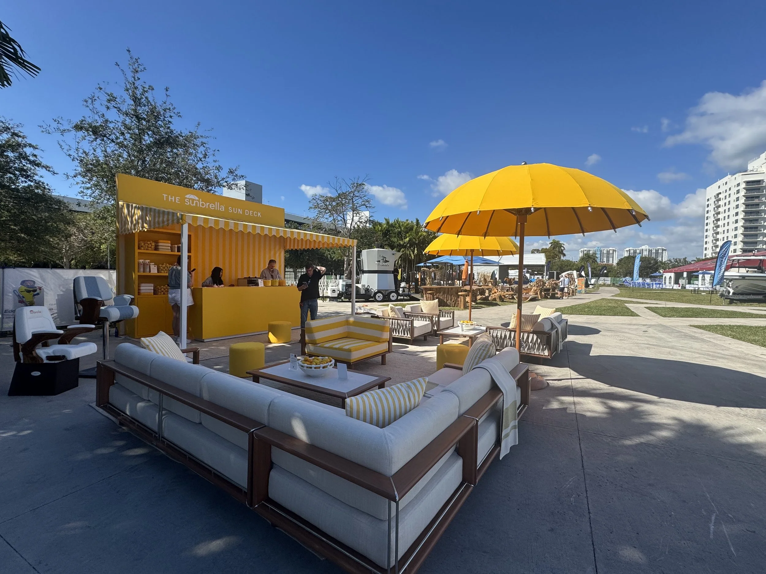 Outdoor patio with yellow umbrellas, white cushioned seating, a yellow kiosk labeled 'The Subrella Sun Deck,' and a clear blue sky with a few clouds. Tall buildings are visible in the background.