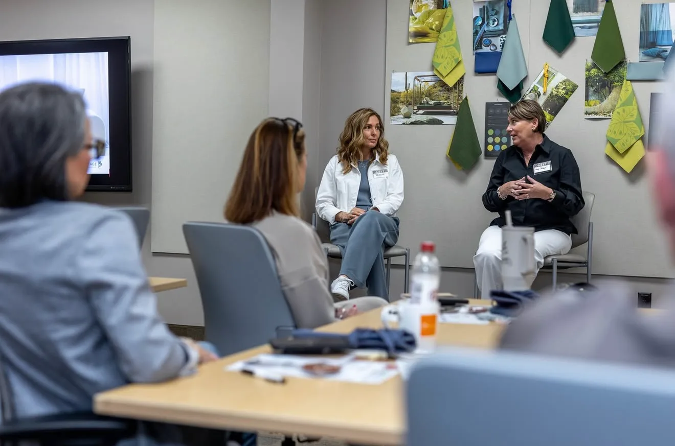 A group of women seated in a meeting room, with two women in the front engaging in a discussion. The room has a wall decorated with fabric swatches and landscape photographs.
