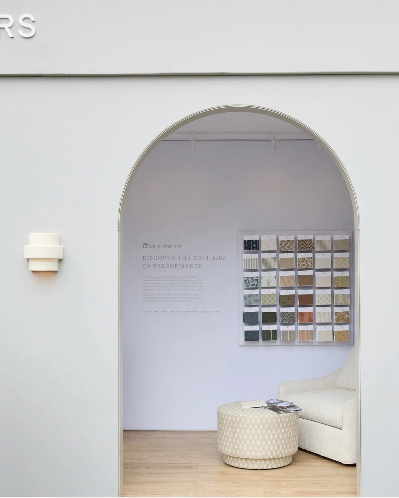 Interior of a modern showroom with a display of fabric samples on the wall and a white armchair with an ottoman nearby.