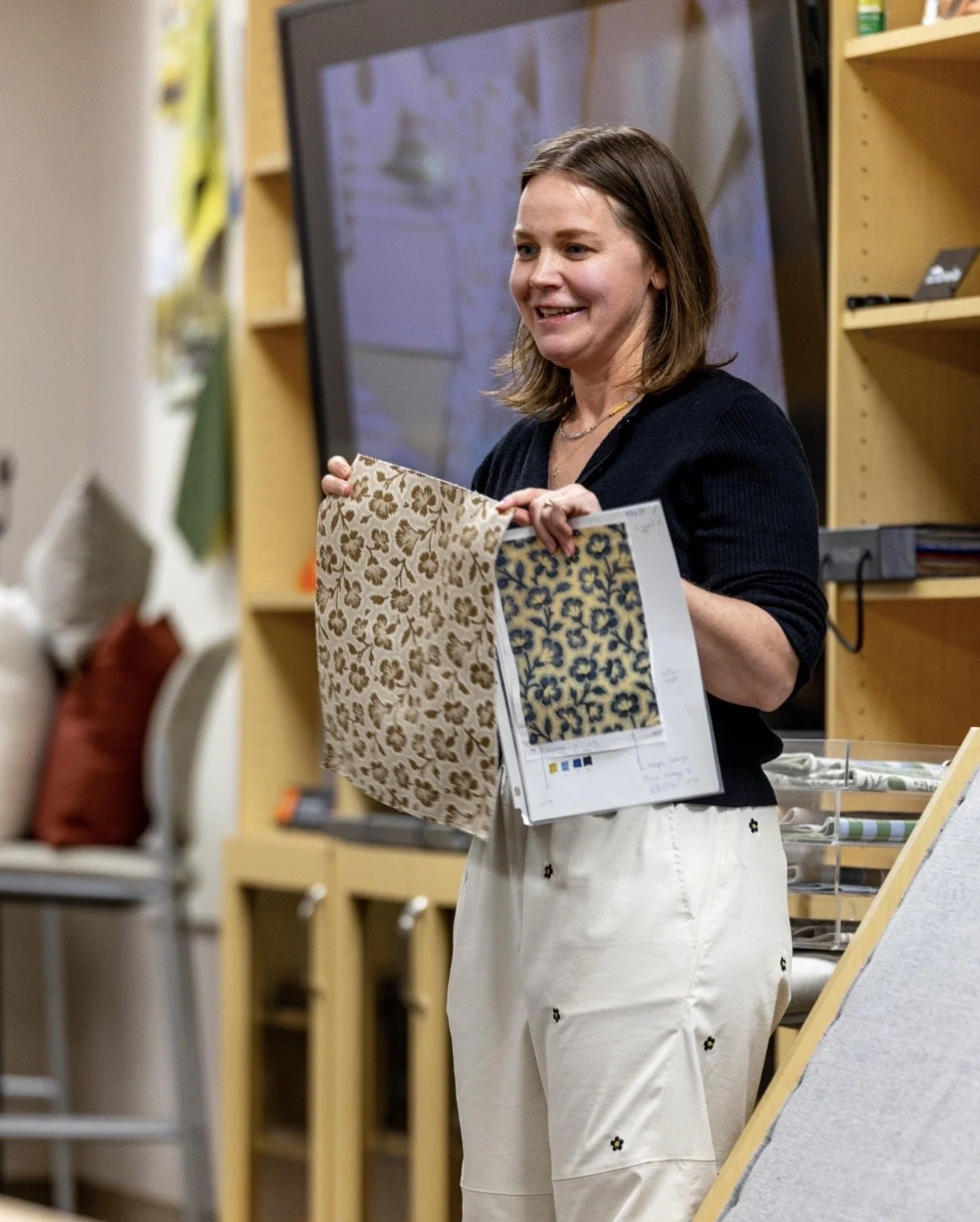A woman is holding fabric samples in a store or showroom, with shelves, a chair, and a large screen or TV in the background.