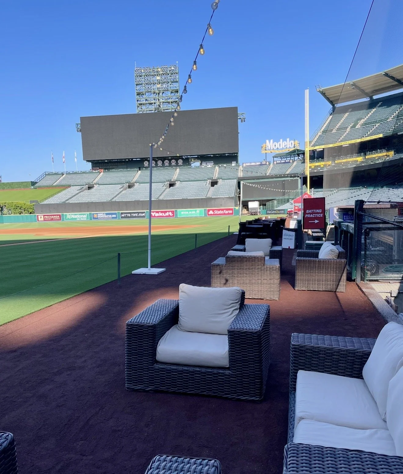 View of an empty baseball stadium with a seating area and a large digital scoreboard in the background, under a clear blue sky.
