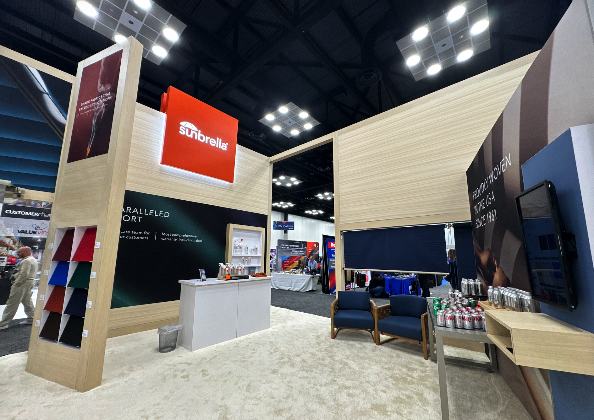 Trade show booth for Sunbrella with fabric swatches on display, seating area with soda cans and water bottles, and promotional banners.
