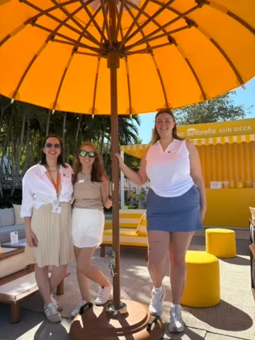 Three women standing under a large yellow umbrella at an outdoor patio area, with seating and a yellow sun deck sign in the background.
