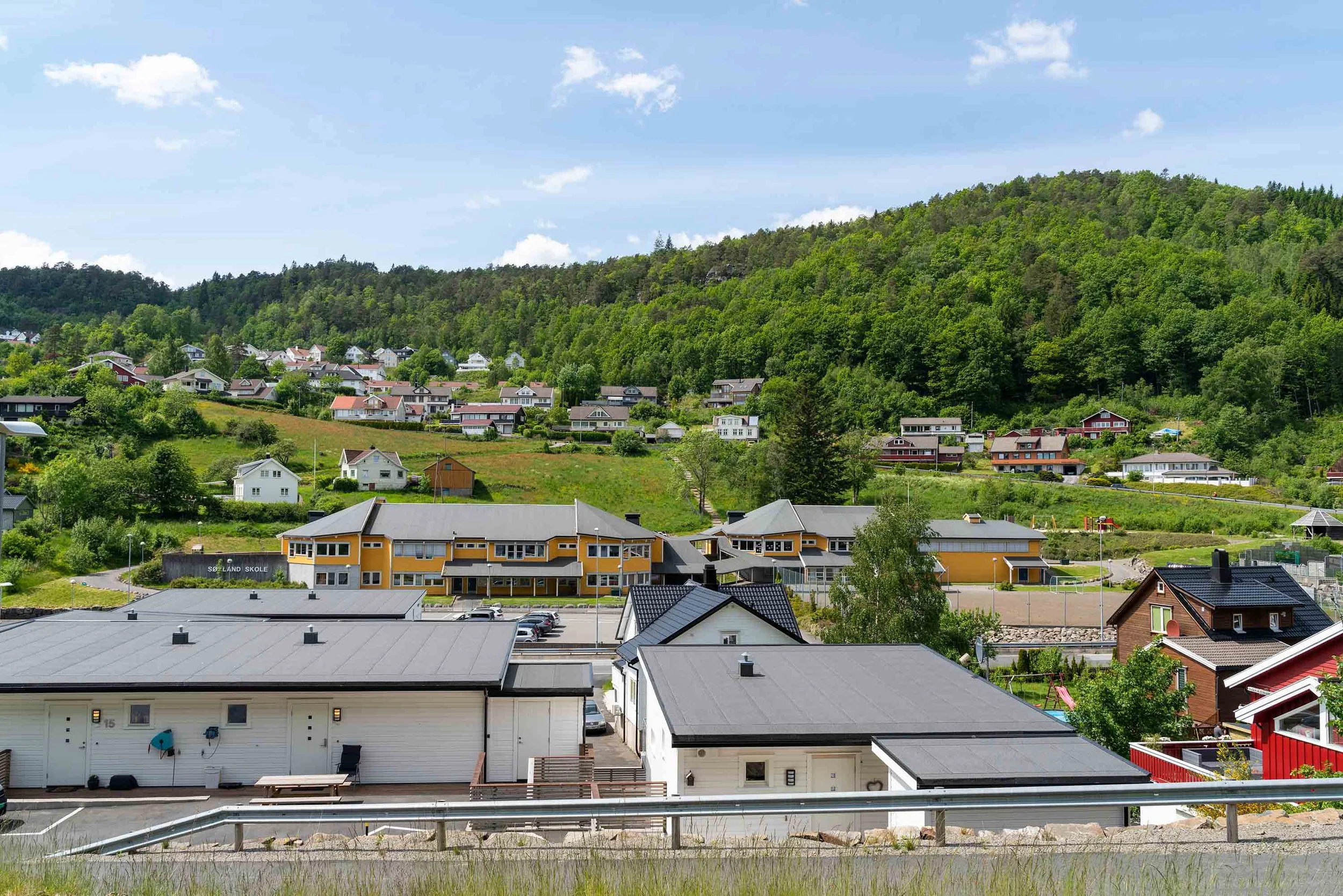 Flerfargede hus i bygata foran en grønn ås med trær, under en skyet blå himmel.