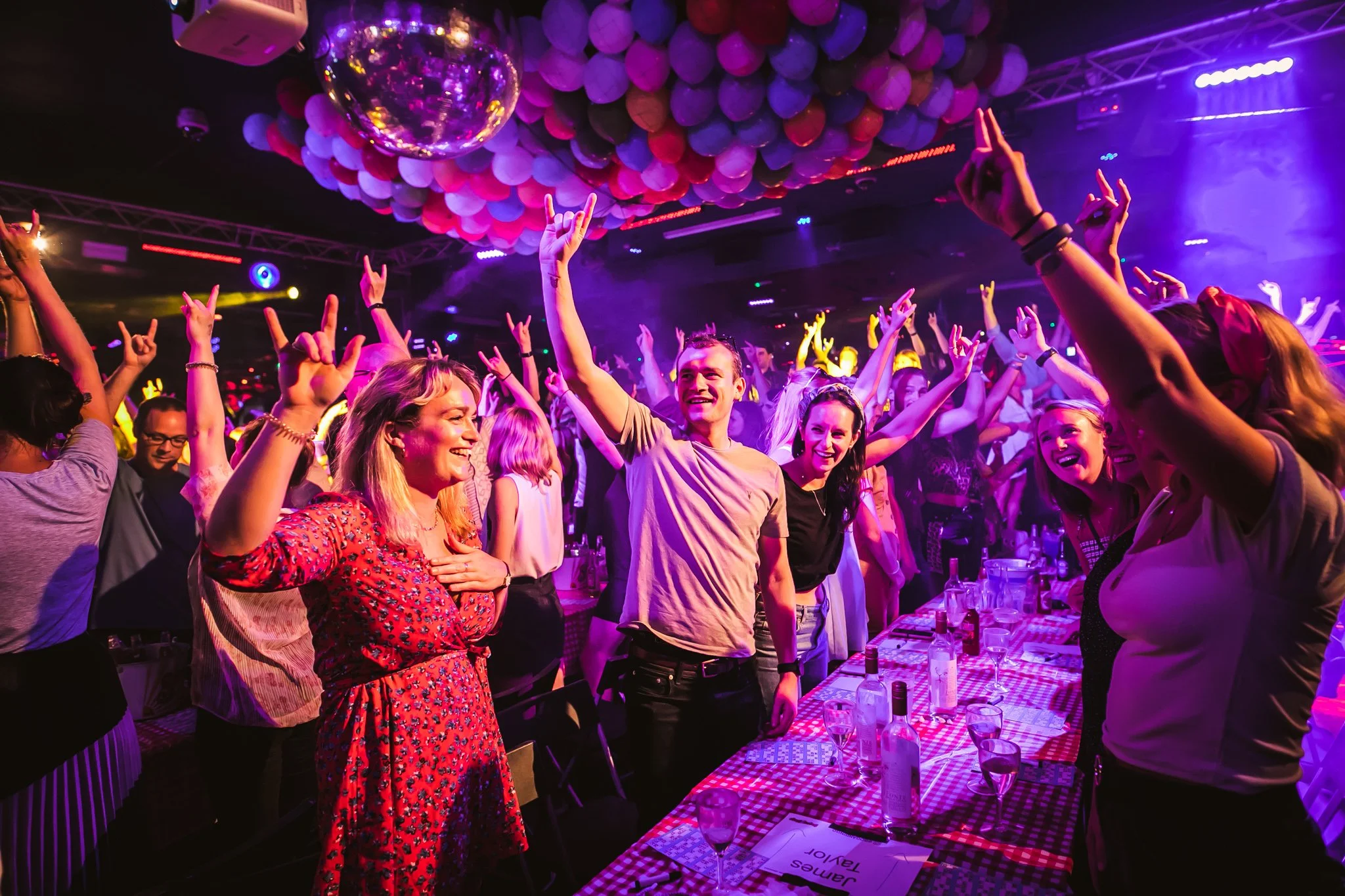 People dancing and enjoying themselves at a nightclub with colorful lighting, balloons hanging from the ceiling, and a disco ball reflecting light.
