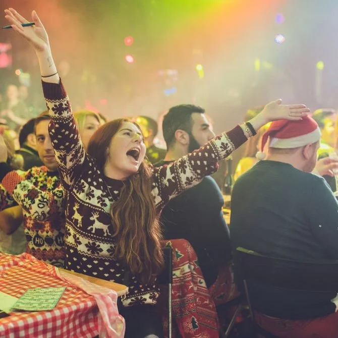 A woman in a Christmas sweater enthusiastically singing or cheering at a holiday party, surrounded by people, some wearing Santa hats, in a festive, decorated setting.