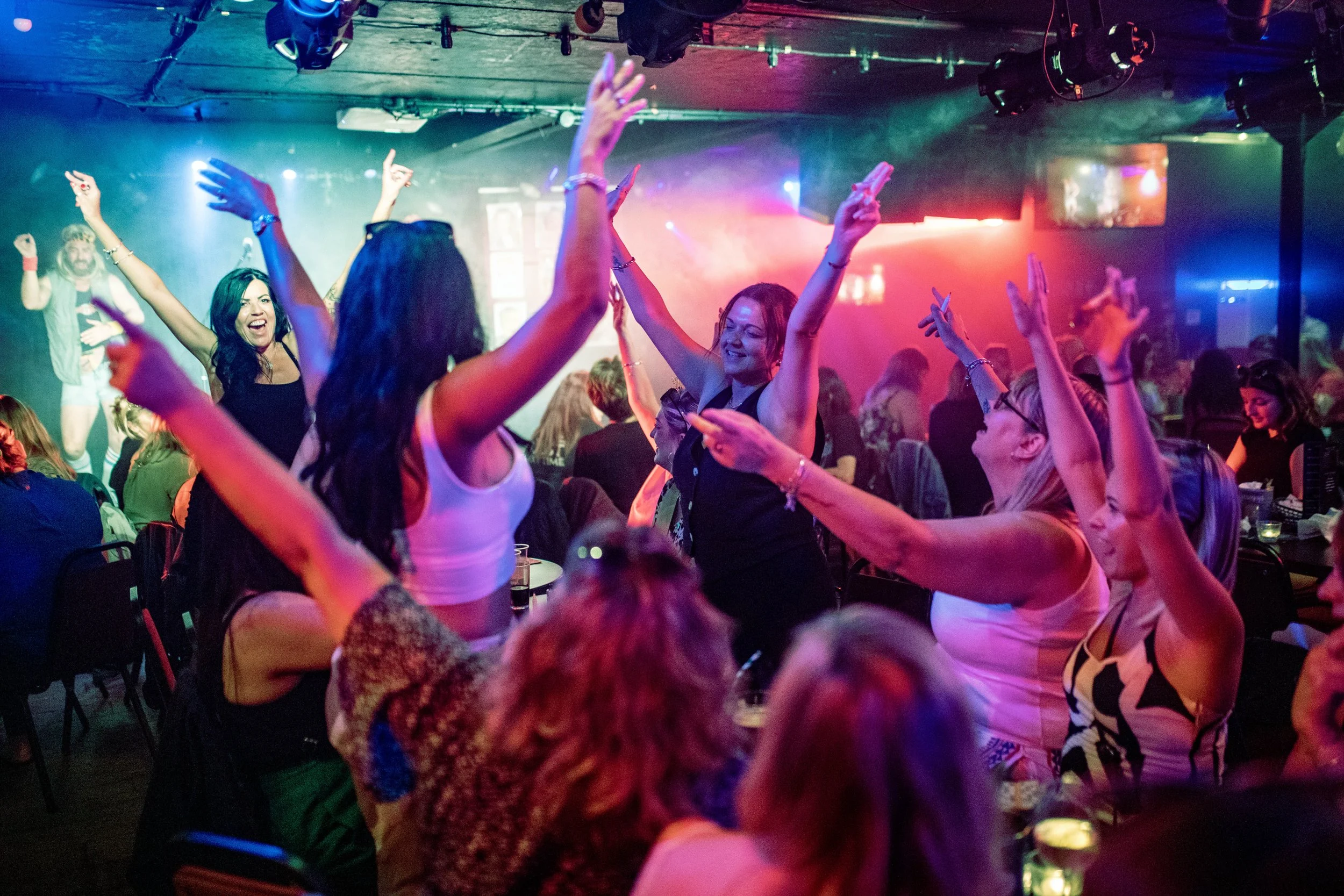 People dancing and enjoying music in a nightclub with colorful lighting and smoke effects.
