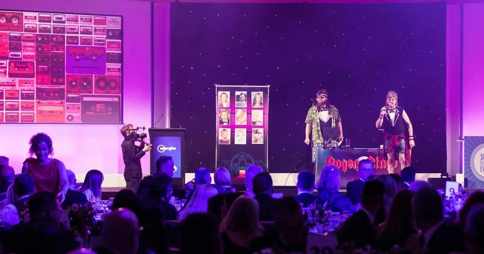 A stage with two performers, one in camouflage shirt and sunglasses, the other in a black tuxedo top with a red plaid shirt tied around the waist, performing at a Rogues Gallery event. An audience watches, and a cameraman records the scene. There are colorful posters, a display of photographs, and a large screen with audio equipment on the stage.