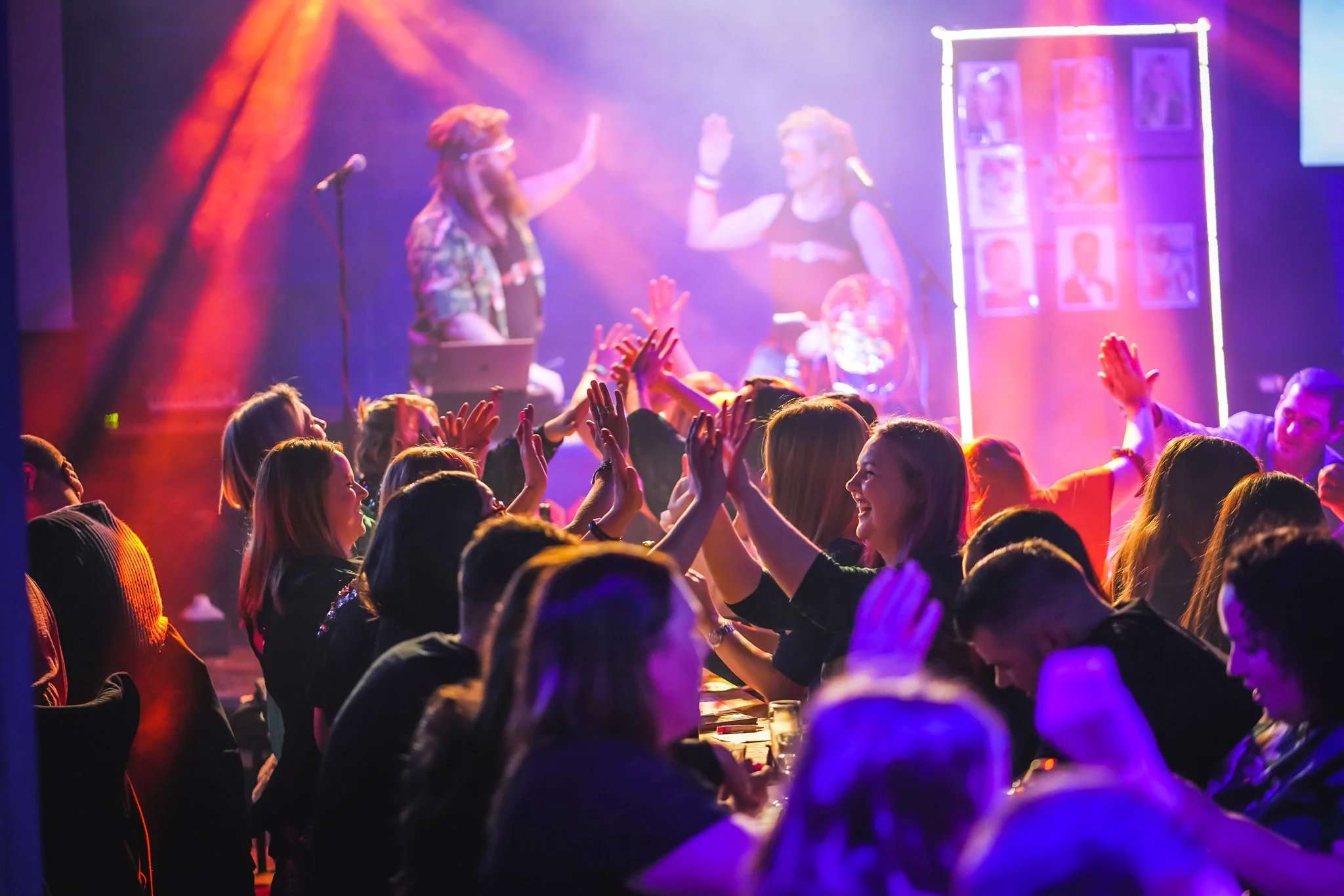 A lively party scene with people dancing and smiling, colorful lighting, and two women high-fiving on stage.