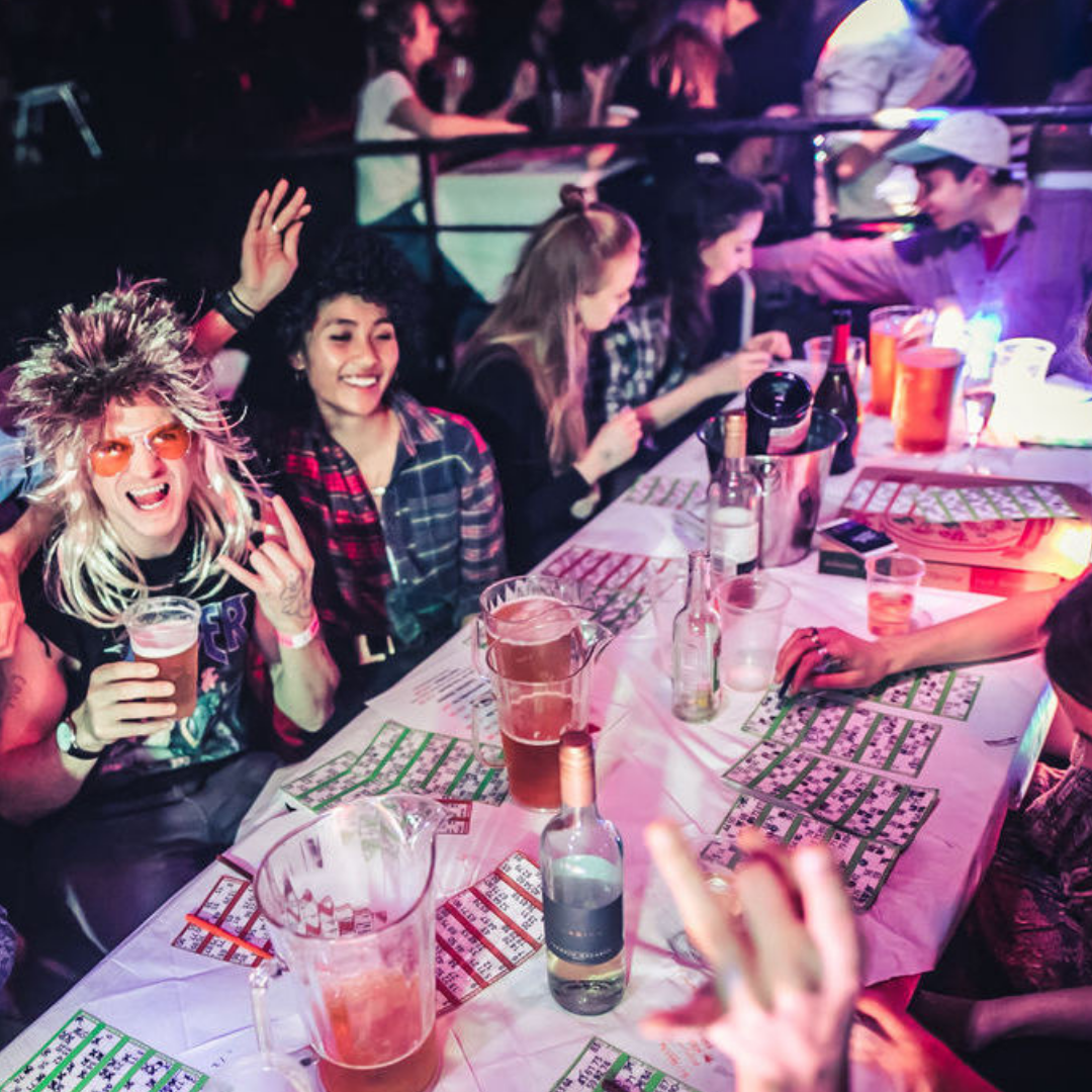Group of people at a party or karaoke event, sitting at a table with drinks, bingo cards, and a cheerful woman with blonde hair and colorful sunglasses, making a rock and roll hand gesture.