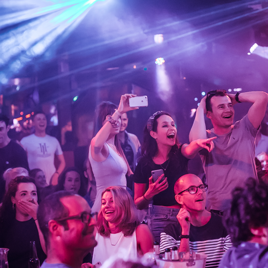 Group of people enjoying live music or performance in a nightclub or concert venue, with colorful stage lights and laser effects.