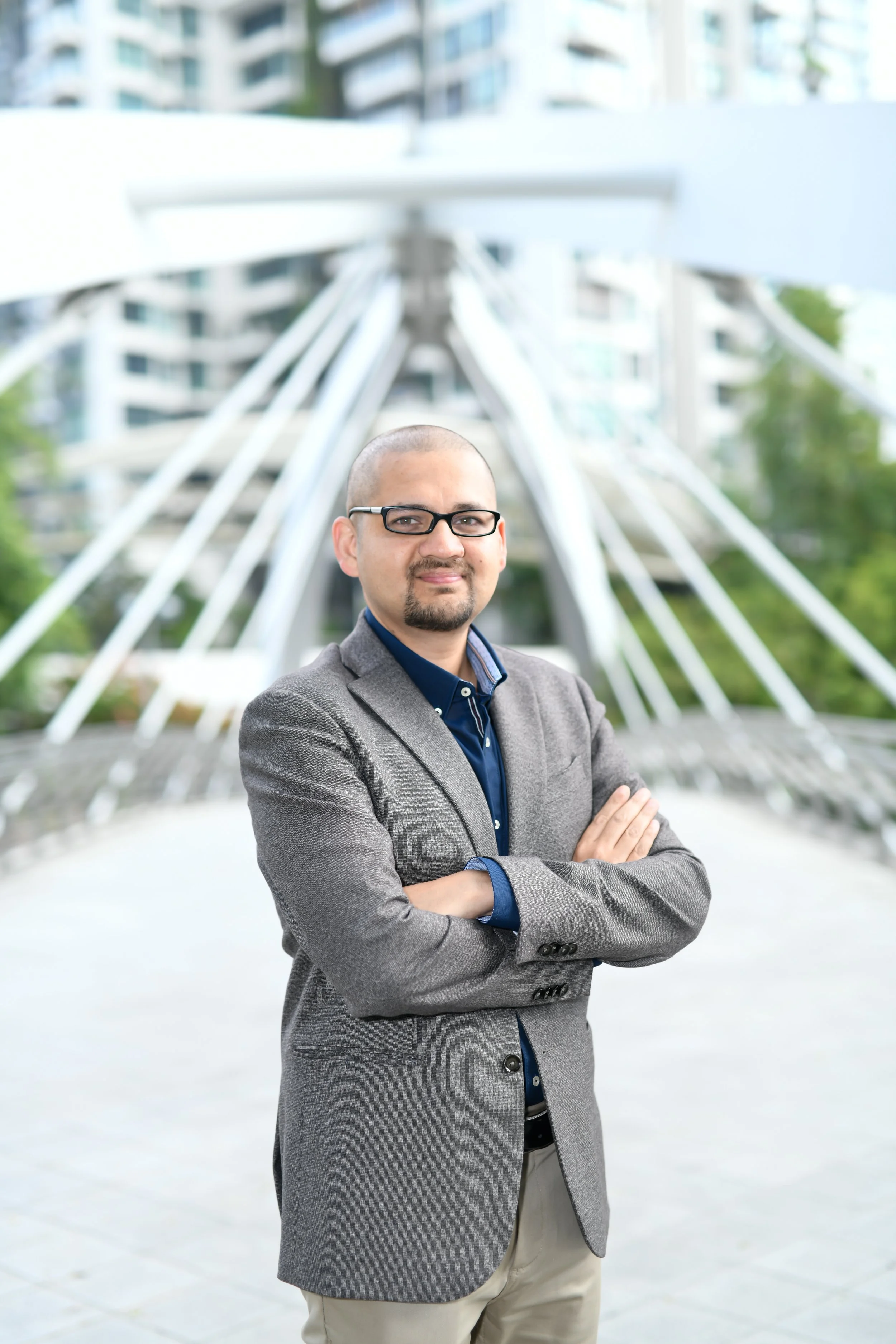 A man with glasses, bald head, and a goatee, dressed in a gray blazer and beige pants, standing outdoors with arms crossed, in front of a modern building with a sculpture that has white beams radiating from a central point.