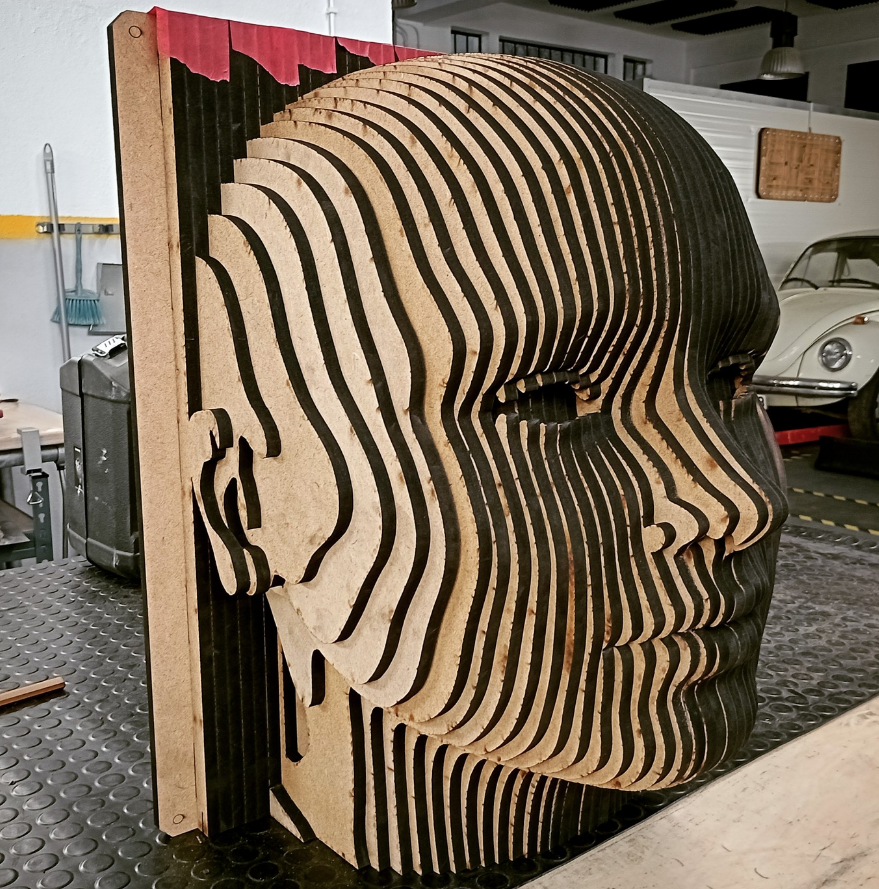 A layered wooden sculpture of a human face in profile view with multiple cutouts creating a 3D effect.