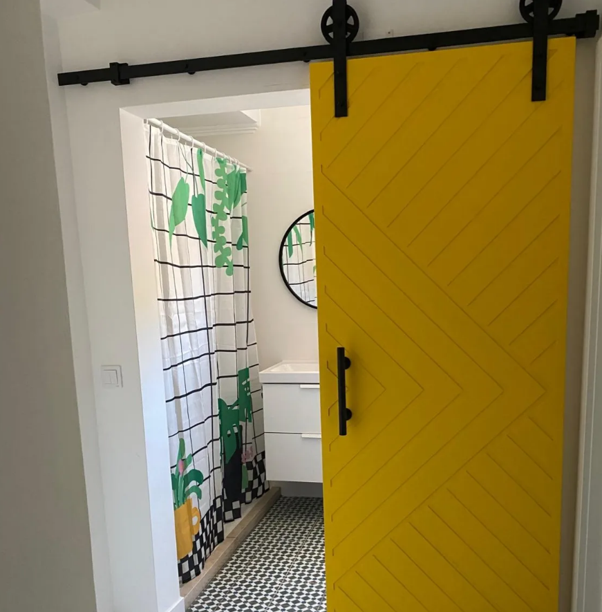 Interior view of a bathroom through a black sliding barn door with a yellow wood panel design. Visible are curtains with a nature and leaf pattern, a white sink vanity, a mirror, and black-and-white patterned floor tiles.