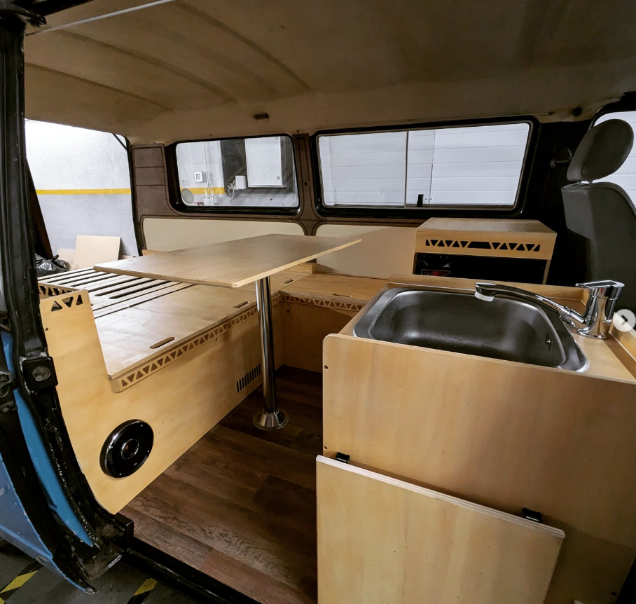 Interior of a converted camper van showing wooden cabinetry, a small sink with a faucet, a foldable table, and windows.