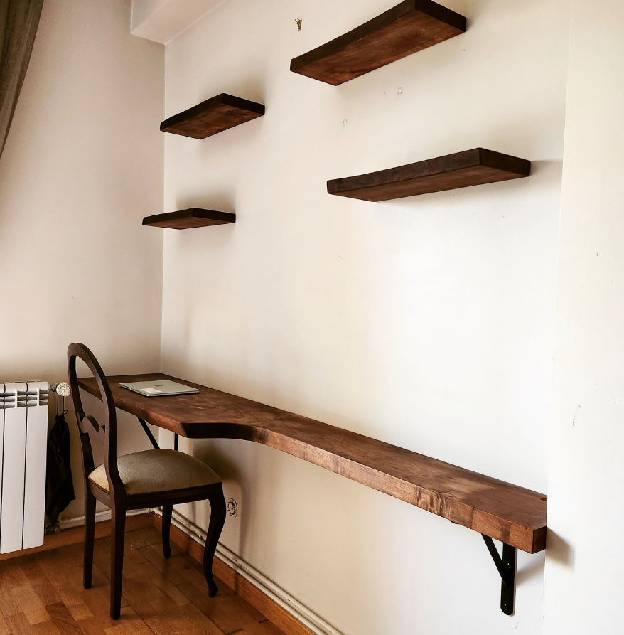 Wall-mounted wooden desk with a chair, three floating shelves on the wall, a laptop, and a radiator.