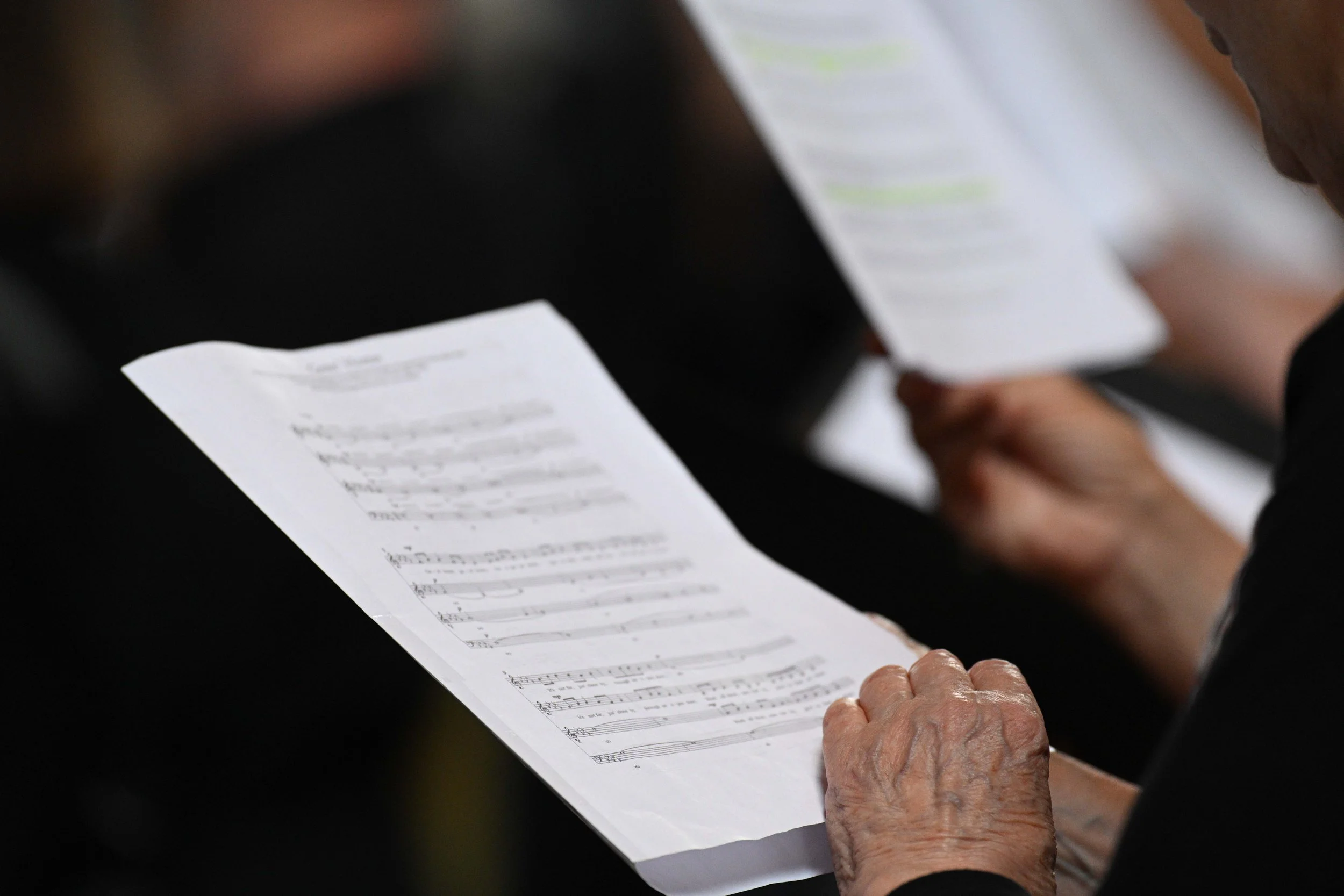 Close-up of elderly person's hand holding a sheet of music, with a younger person's hand also visible holding the same sheet, in a music setting.