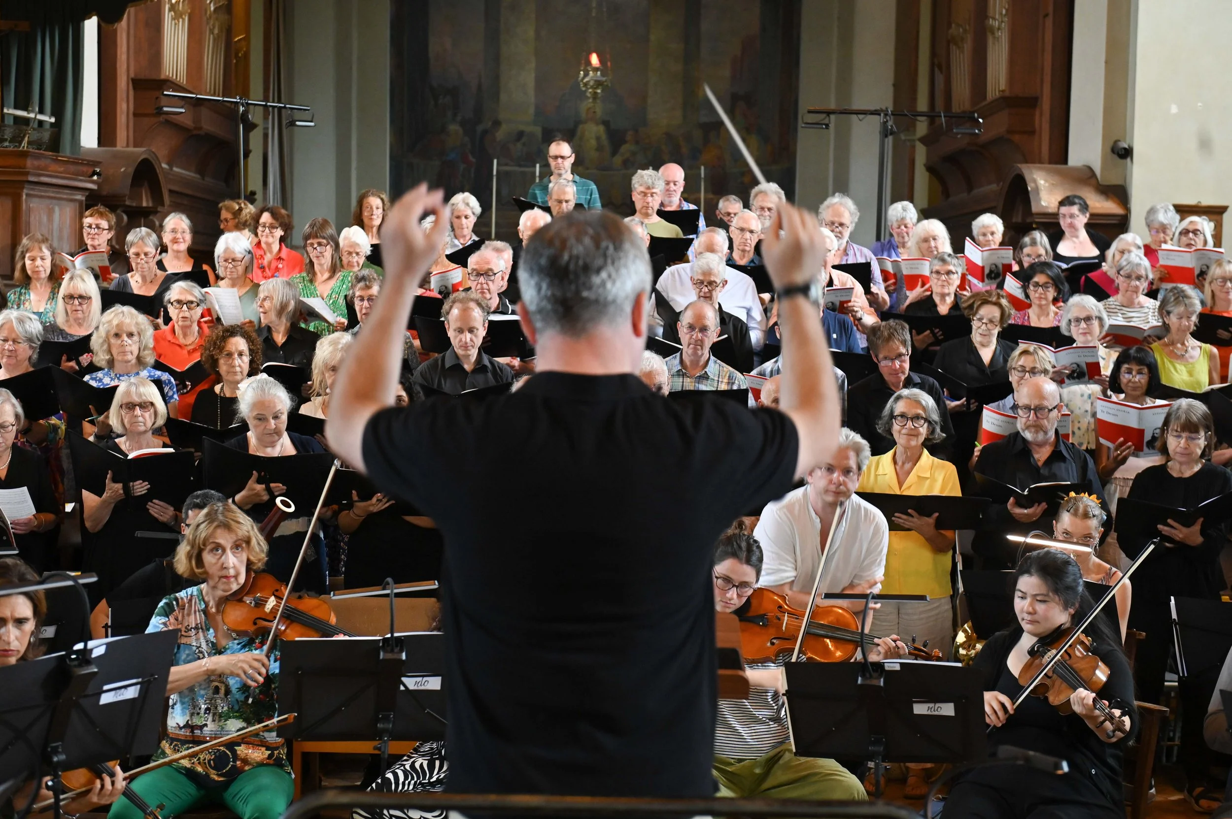 A choir and orchestra in a concert hall with a conductor leading them. The piano and violins are visible, and the audience is not visible.