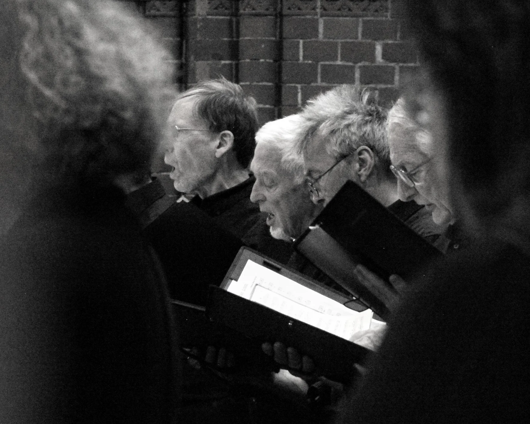 Four people sitting closely together at a table in a dimly lit room, looking at open books or documents. The background features a brick wall, indicating an indoor setting, possibly a study or a meeting space.