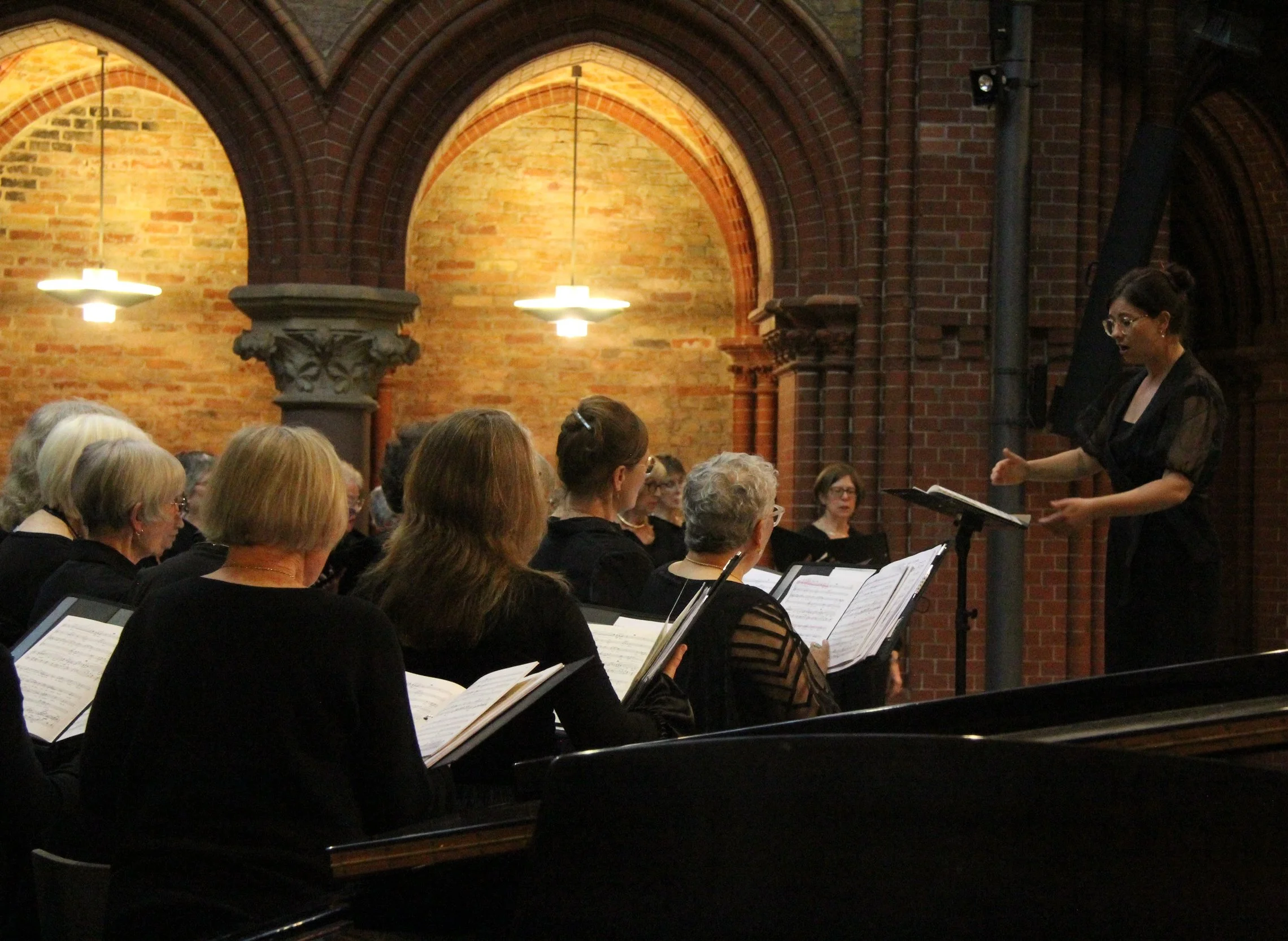A woman is conducting a choir of women singing in a room with arched brick walls and hanging lights.