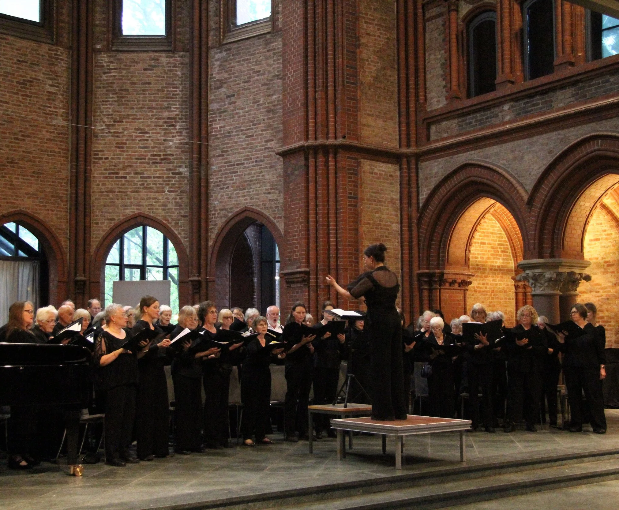 A choir performing in a large church with brick walls and tall arched windows, with a conductor standing in front of them.
