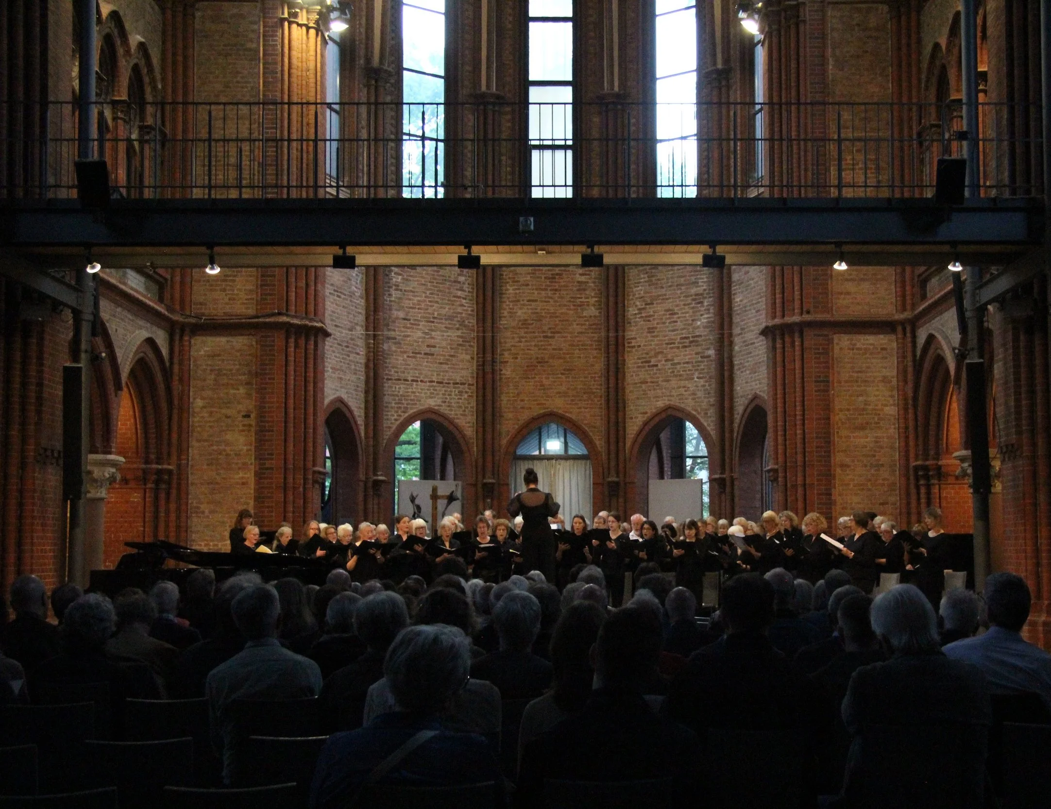 A choir performing in a church with arched windows, brick walls, and a large audience watching.