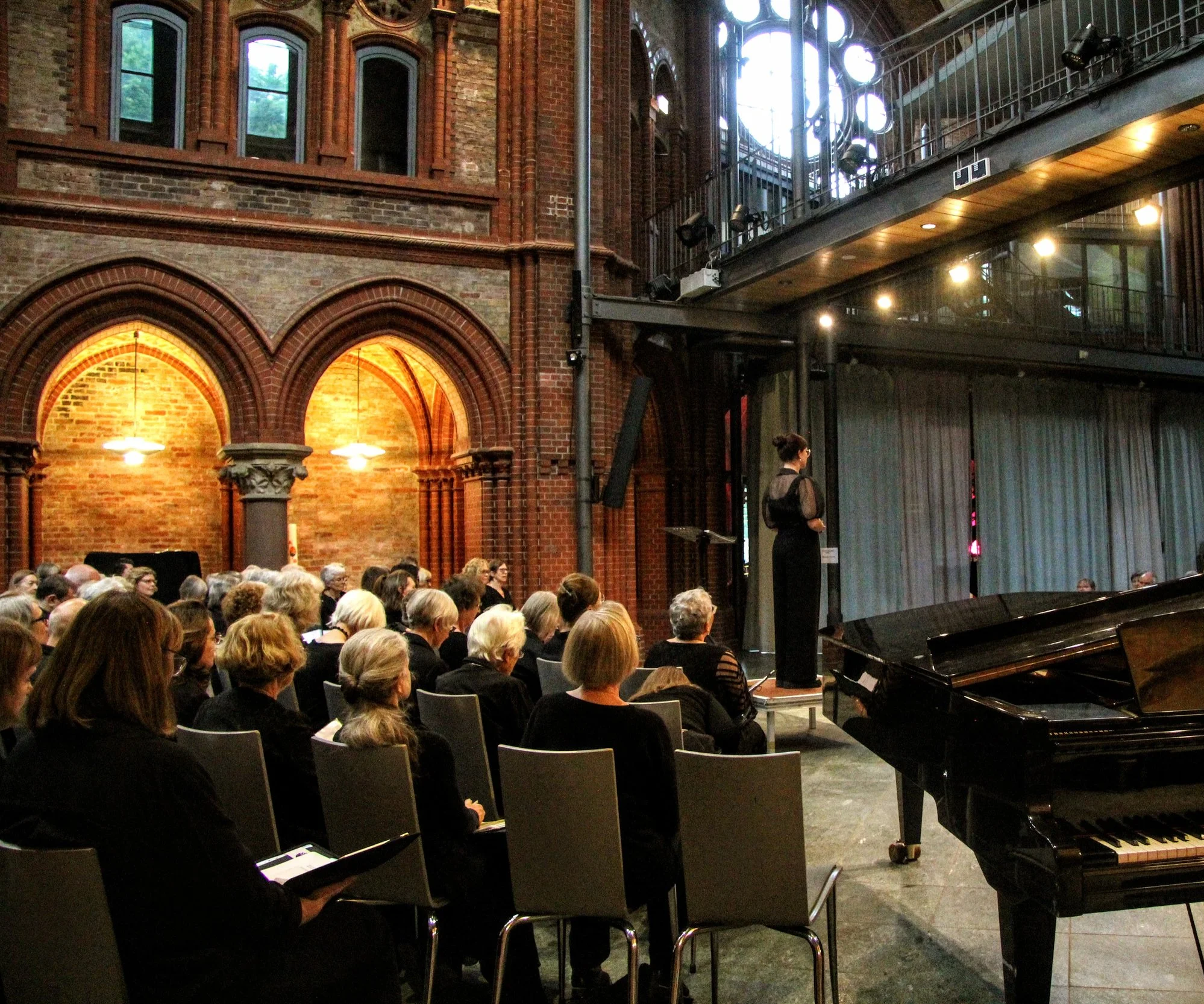 An indoor concert hall with a classical brick architecture, filled with an audience seated in rows, a performer playing the piano on stage, and a woman standing near the piano. The hall features tall windows and modern lighting fixtures.
