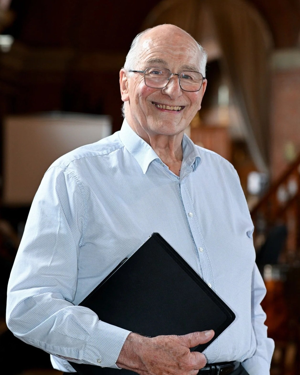 An elderly man with glasses, smiling, holding a closed black folder or notebook, standing indoors with a blurred wooden background.
