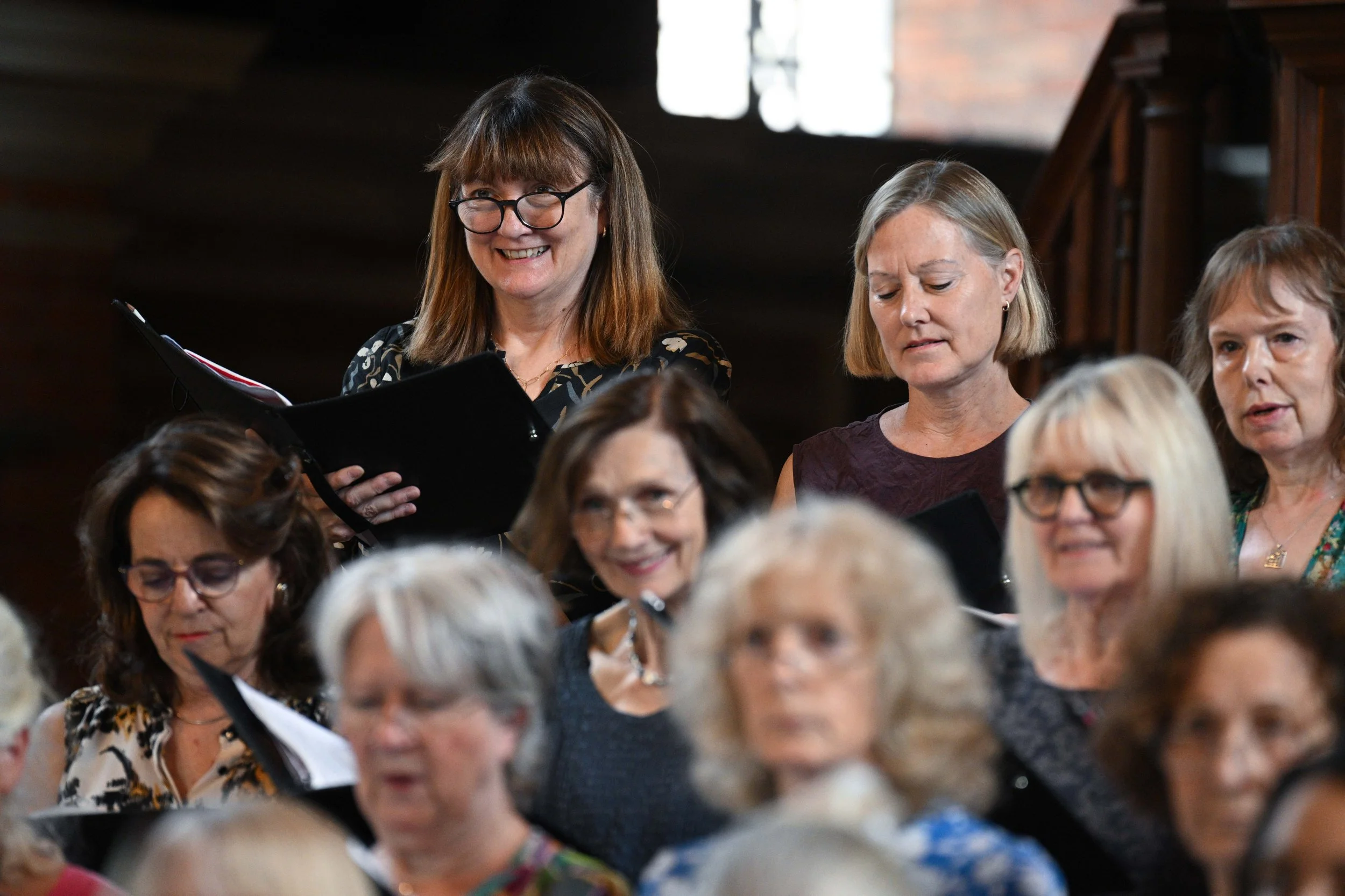 A group of middle-aged women singing together, some holding songbooks, in a cozy indoor setting with warm lighting and wooden details.