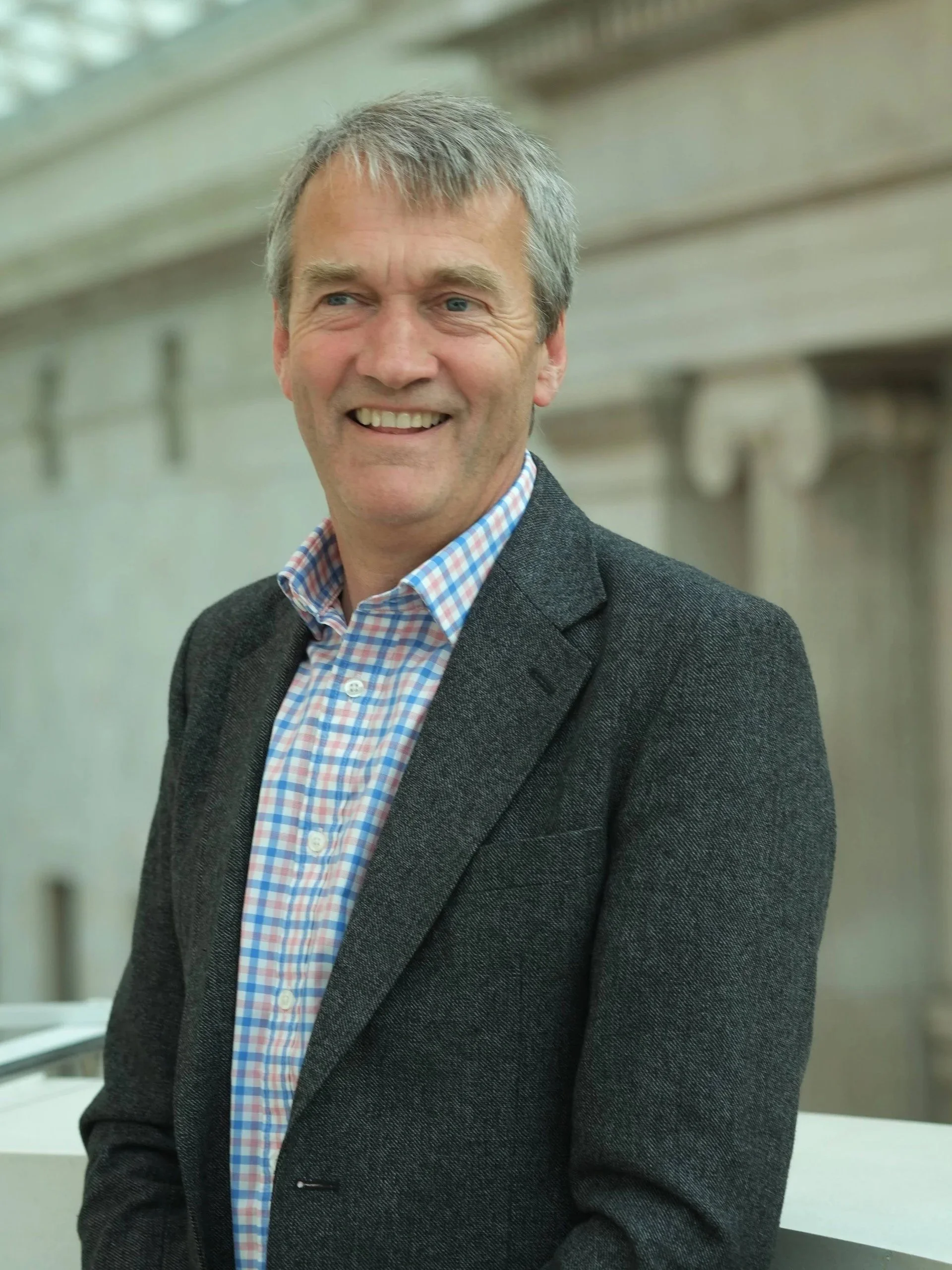 A middle-aged man with gray hair and a light complexion, smiling, wearing a dark blazer and a blue and white checkered shirt, standing indoors with a modern background.
