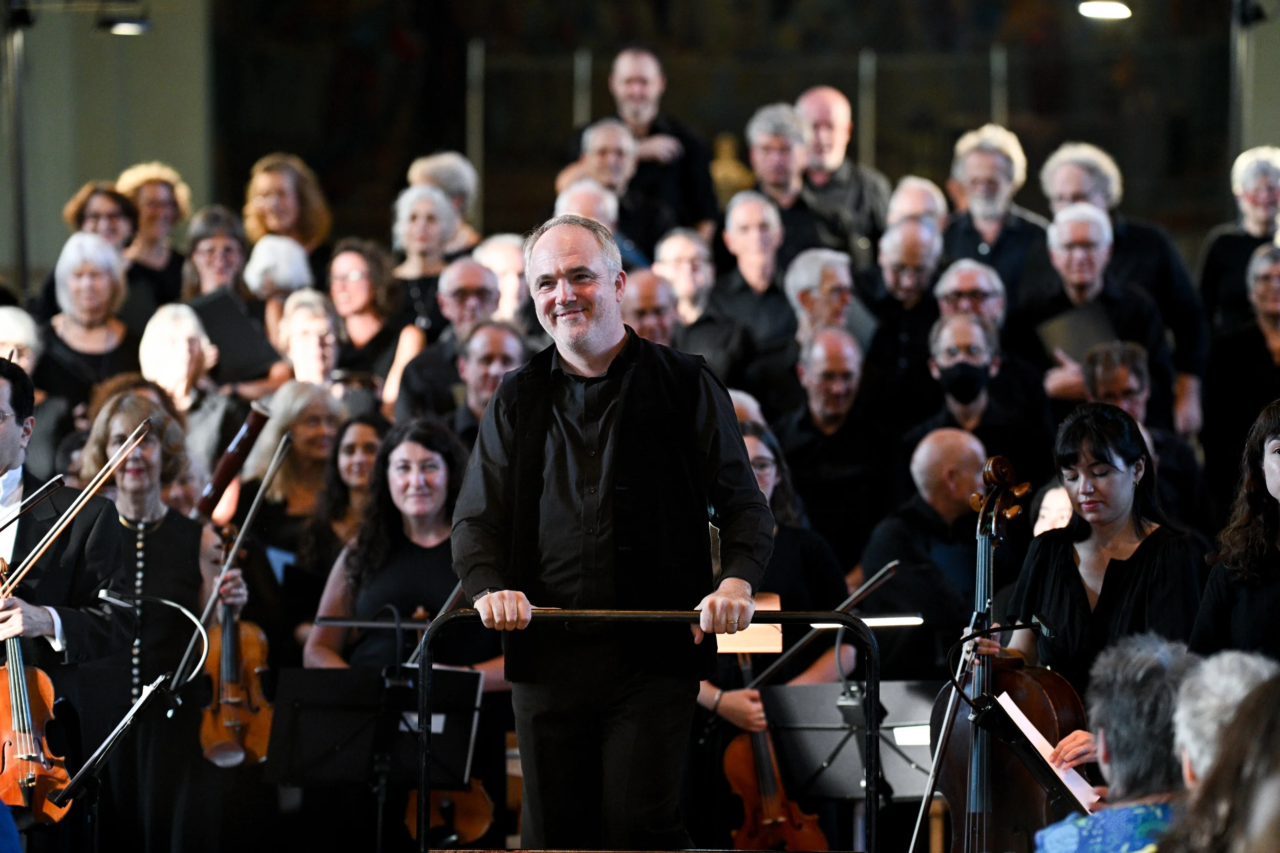 A male conductor leading an orchestra with many musicians, many of whom are playing string instruments, in a concert hall.