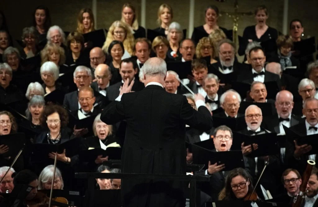 A large choir singing with a conductor leading them, all dressed in formal black attire, during a performance.