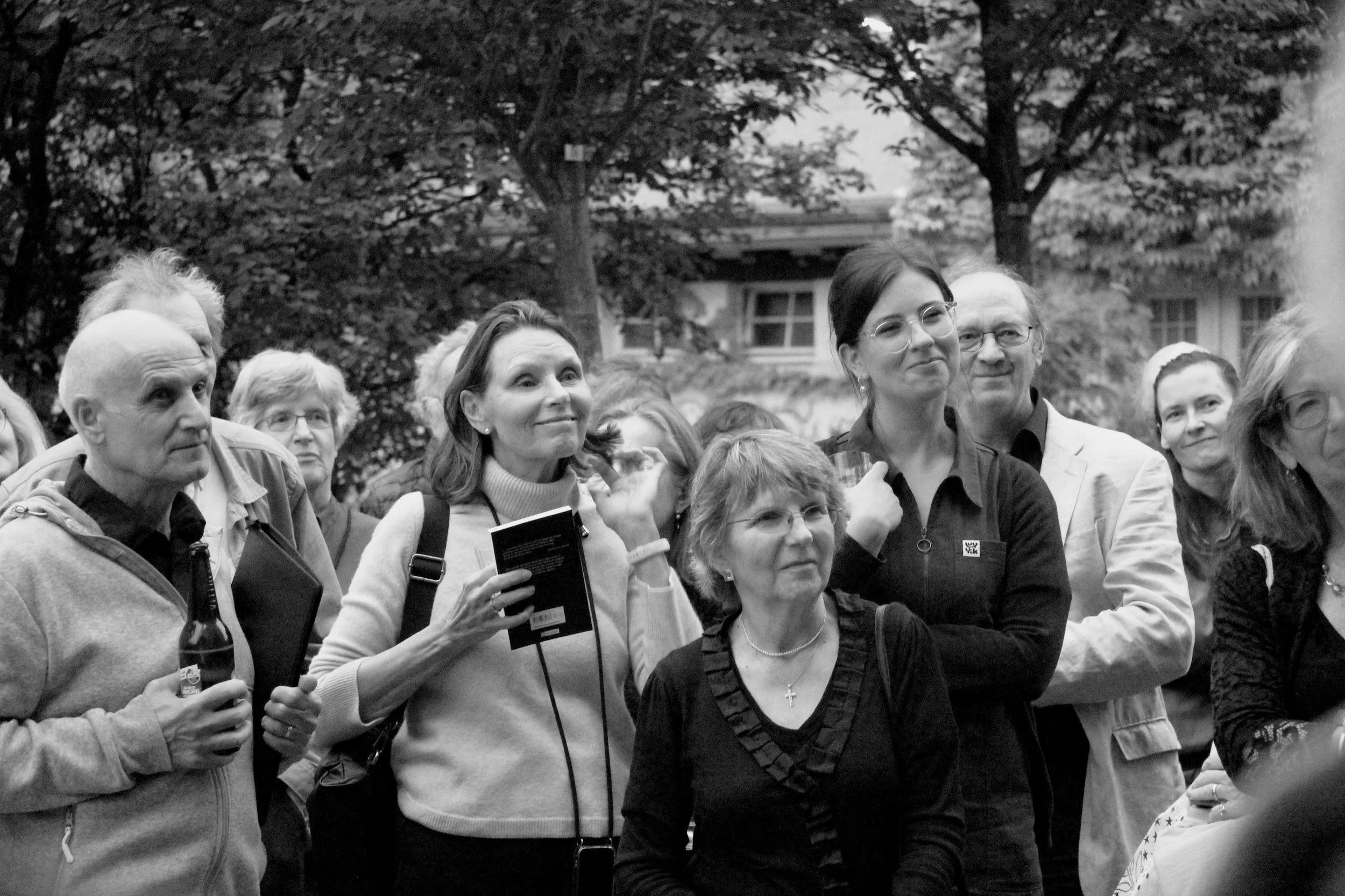 A group of people attending an outdoor event, with trees in the background. They are engaged, some smiling, and holding items like a booklet and drinks.